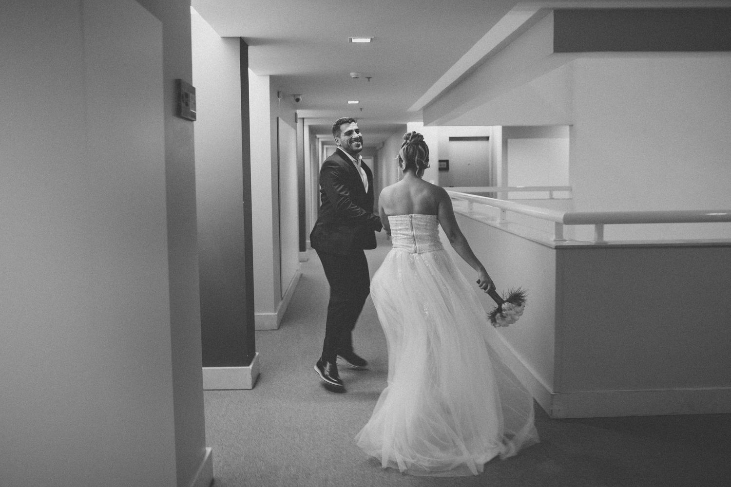 A couple dances and laughs together in a hallway wearing wedding attire.