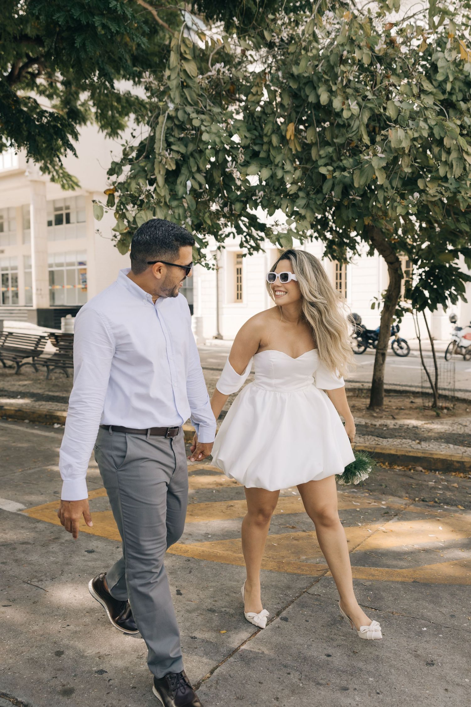 A couple walks together under a tree canopy, with the bride wearing a short white dress and sunglasses.