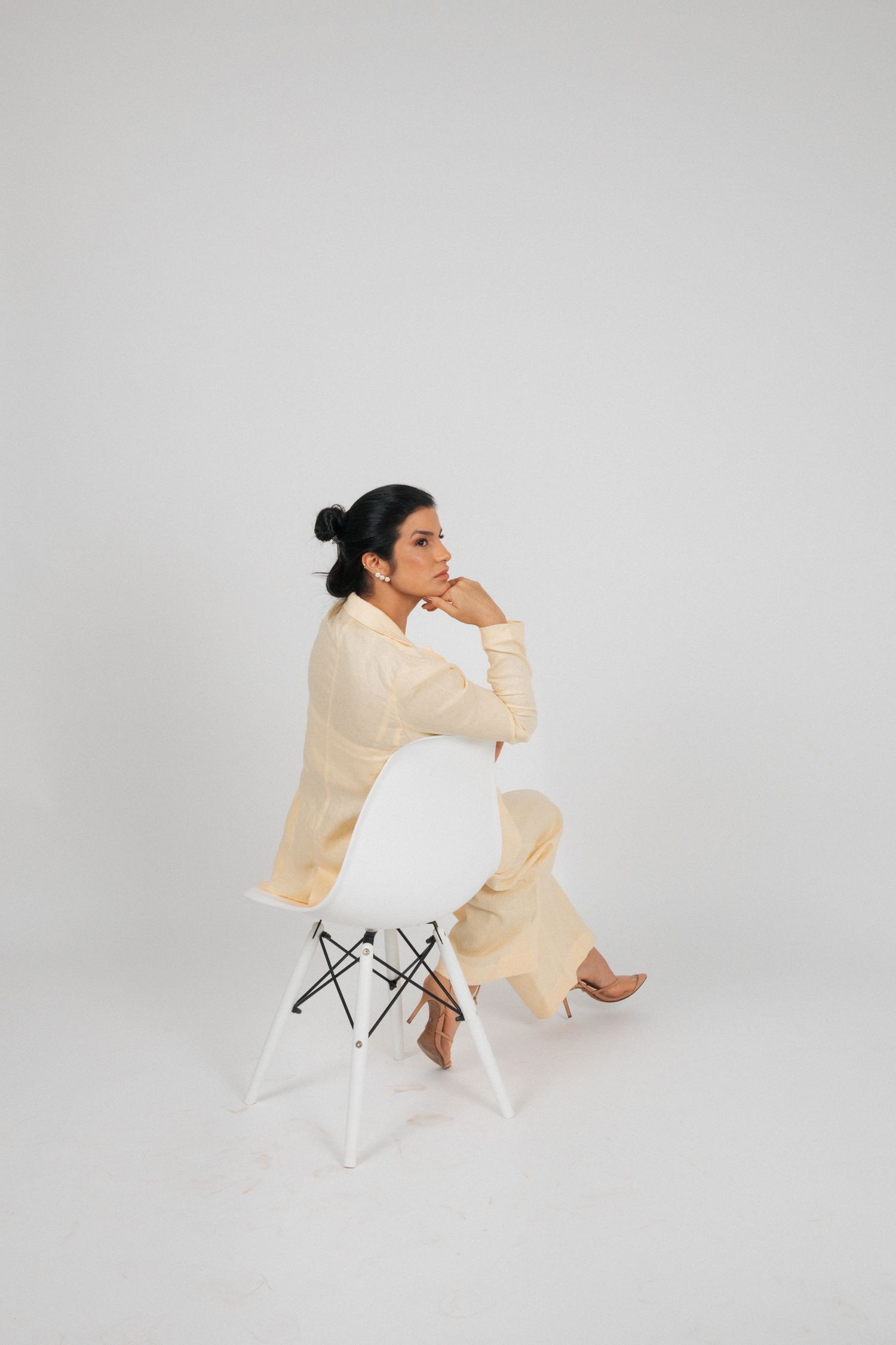 Person in cream colored outfit sitting thoughtfully on a white designer chair against light gray backdrop.