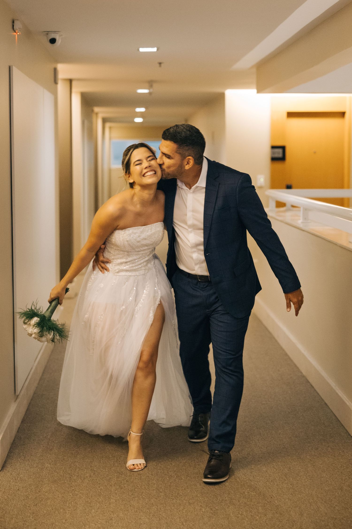 A joyful couple walks through a hotel hallway in formal attire.