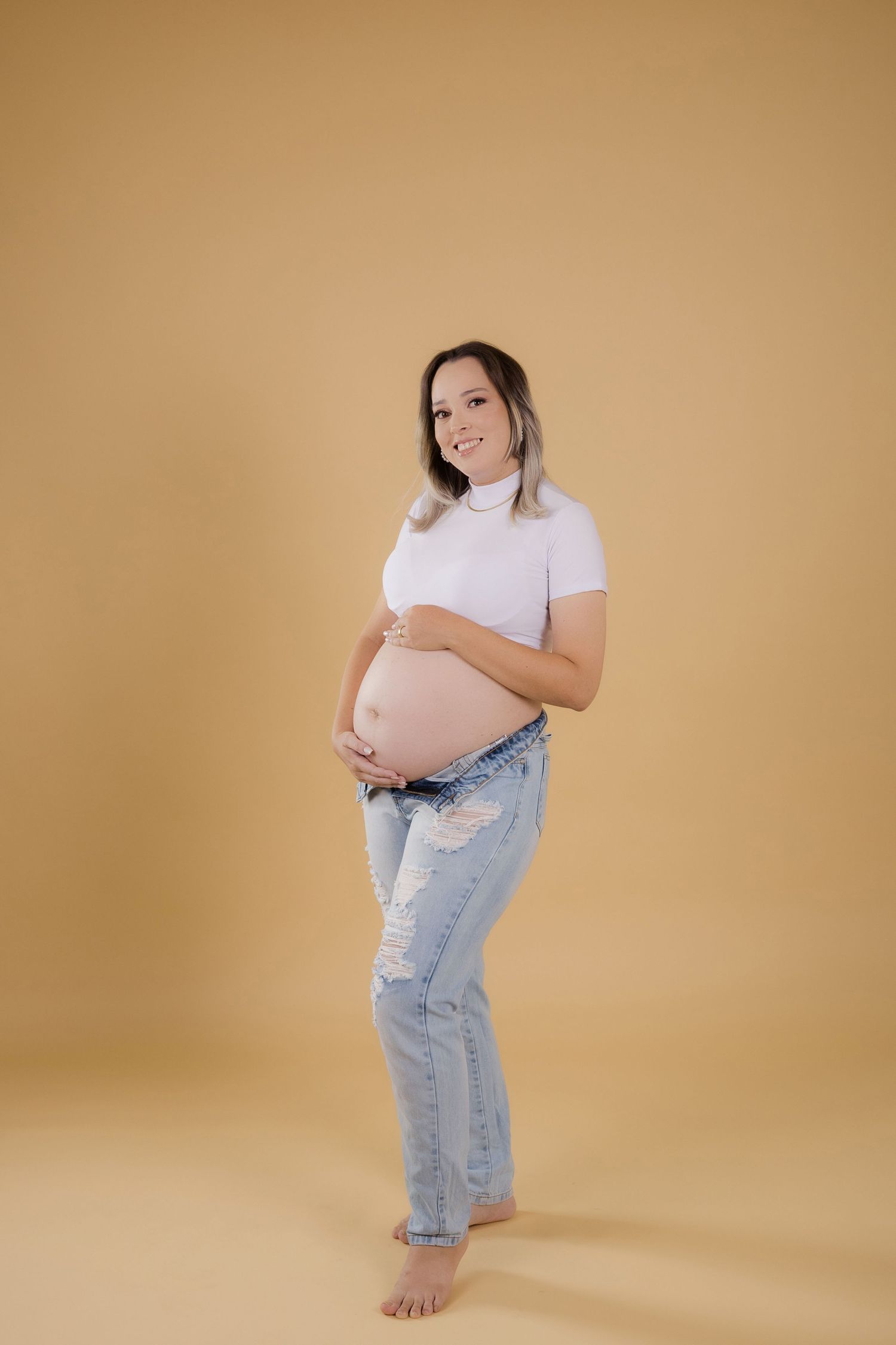 Expectant woman in white t-shirt and distressed jeans poses against beige backdrop during maternity photoshoot.