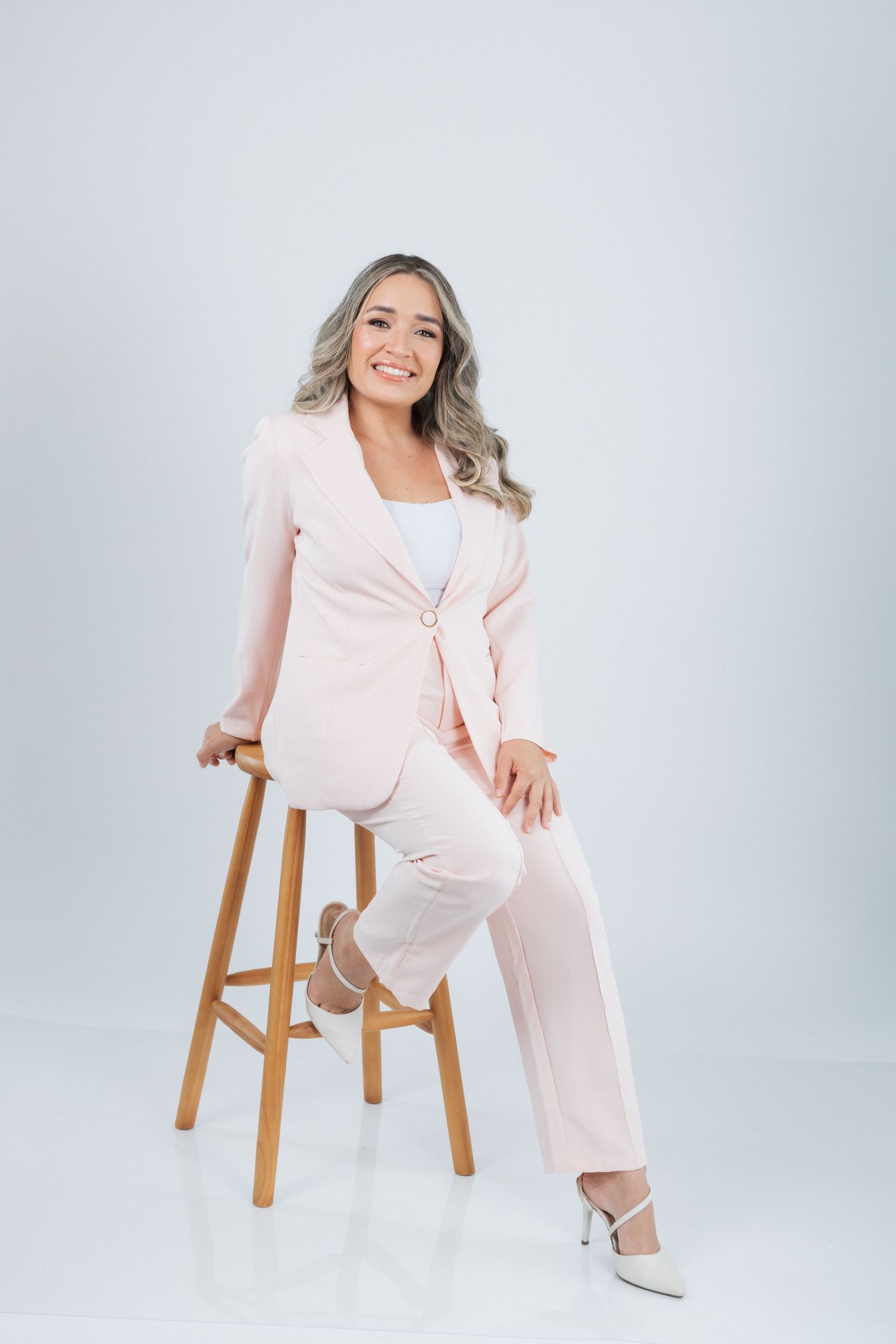 Professional wearing a light pink business suit and white top sits elegantly on a wooden stool against a gray background.