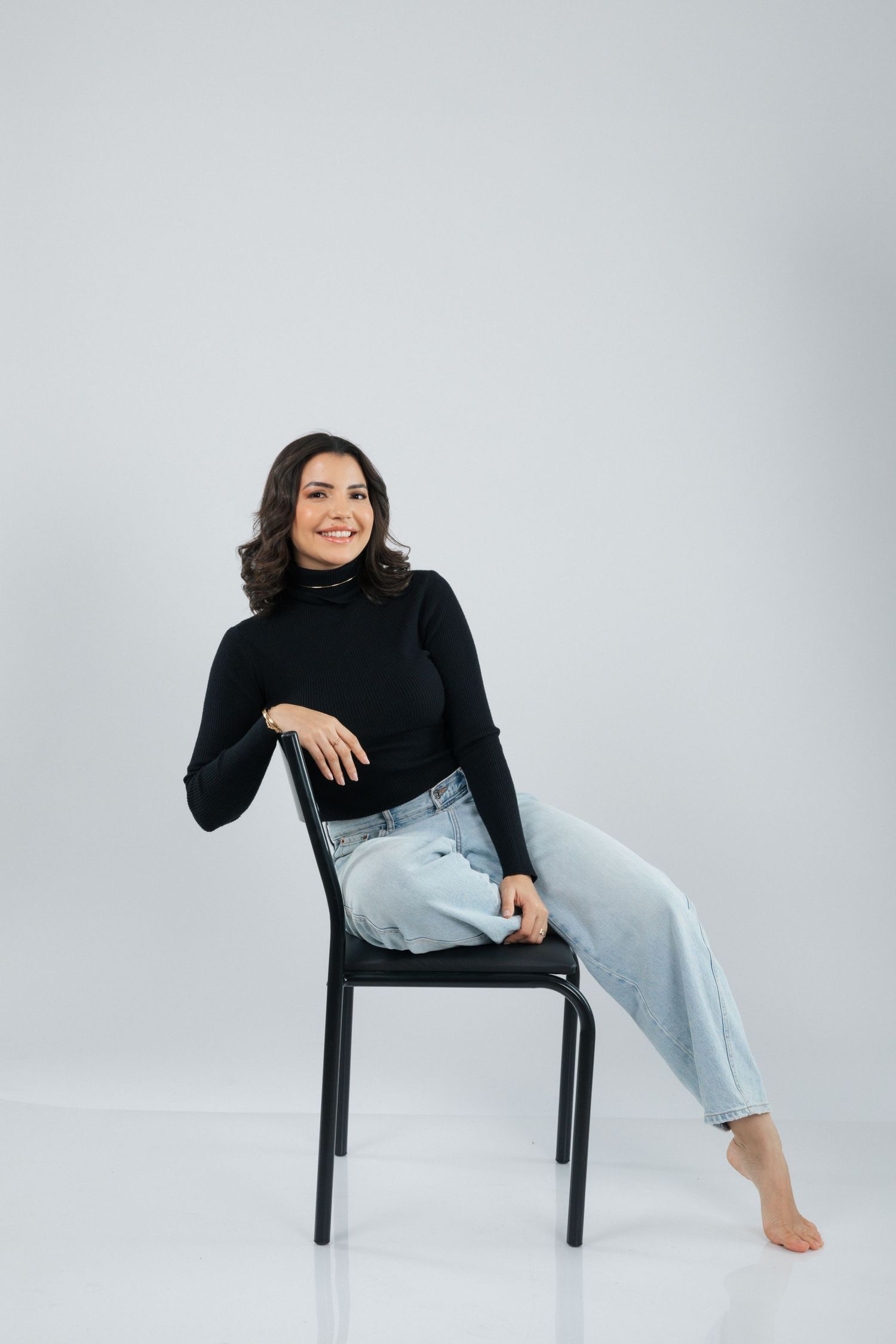 Model relaxes in casual pose on black chair wearing black sweater and light jeans.