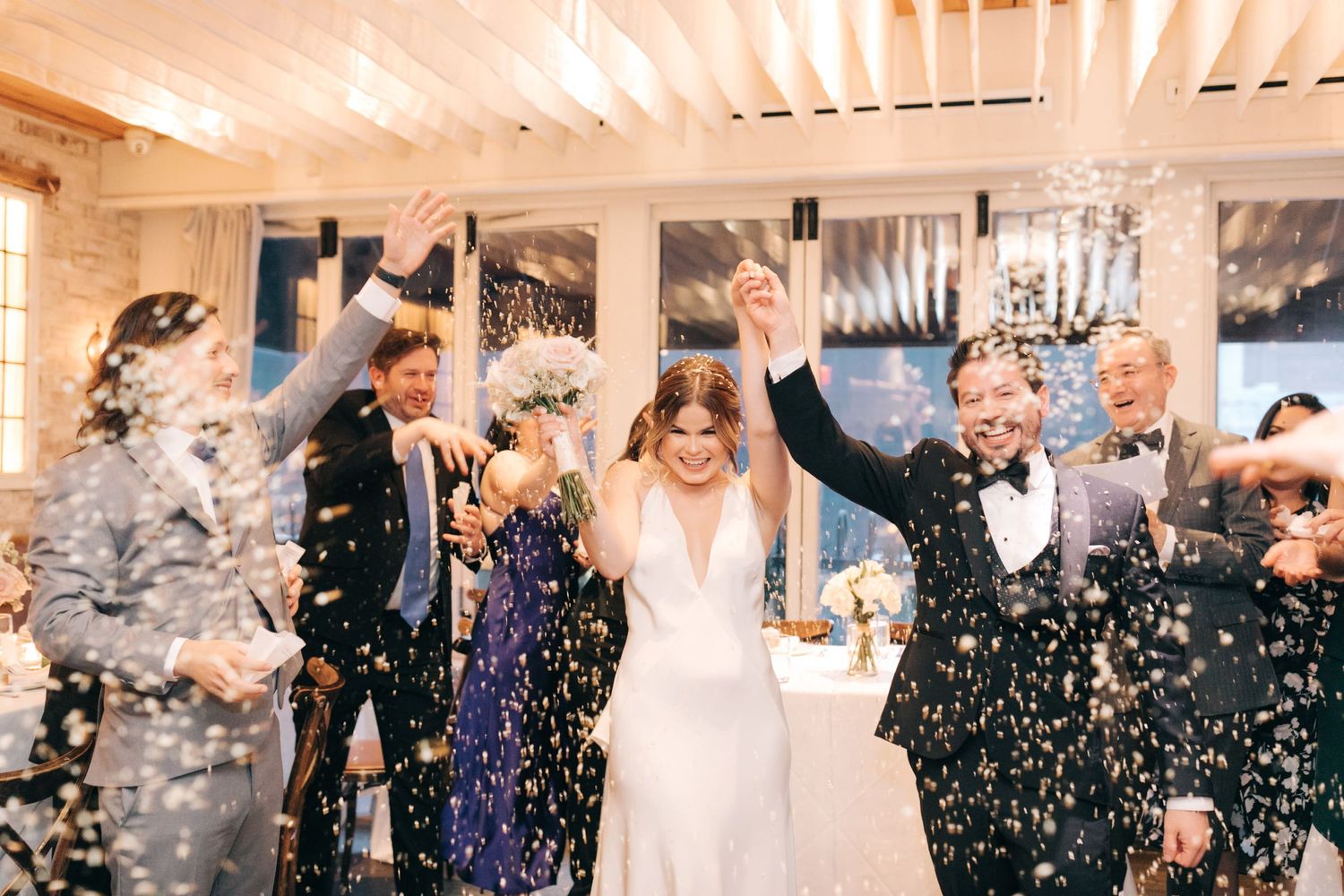 Wedding celebration with people cheering as confetti rains down in an elegant indoor reception venue.