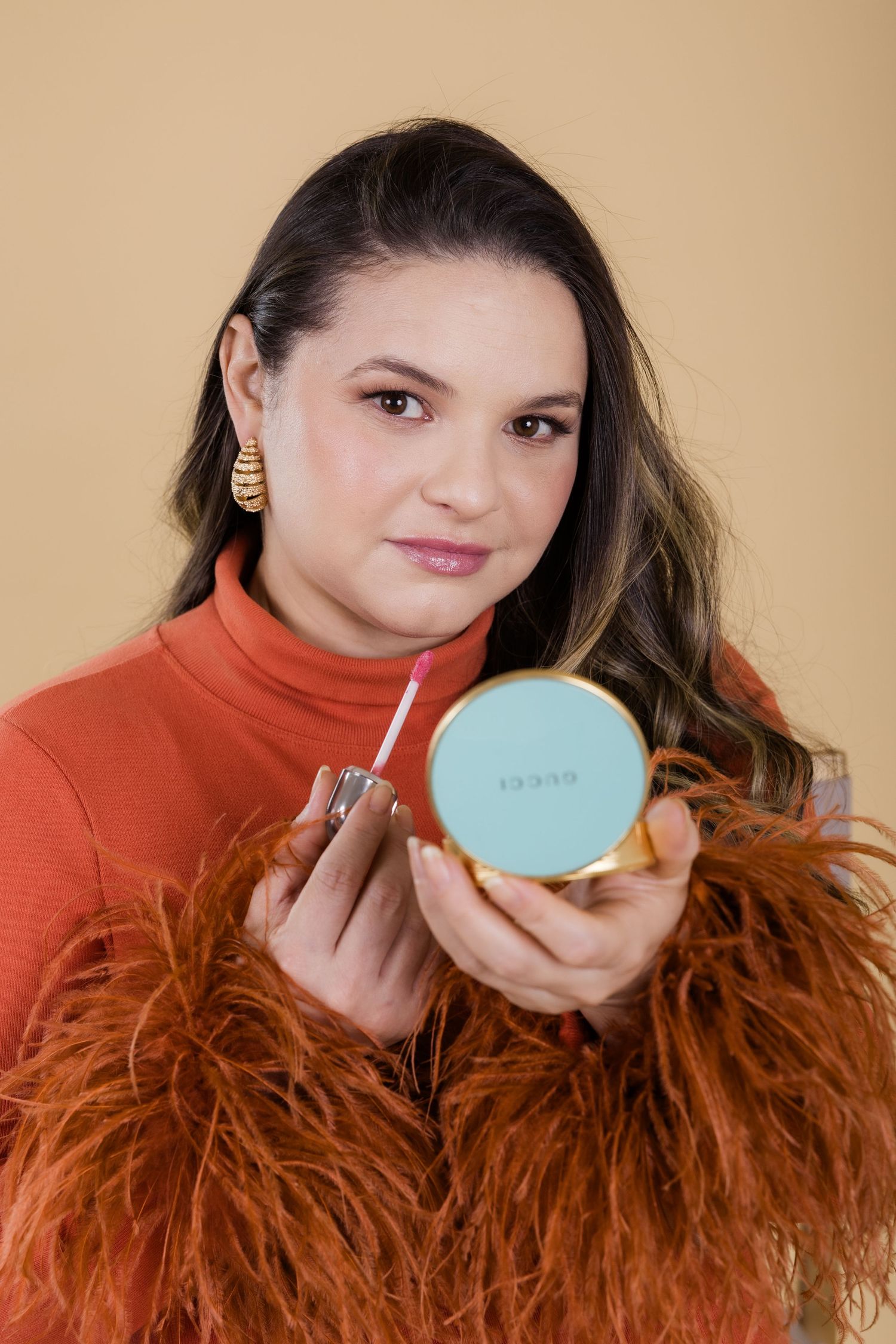A close-up shot shows someone holding a turquoise compact while wearing an orange turtleneck and feathered accessories.