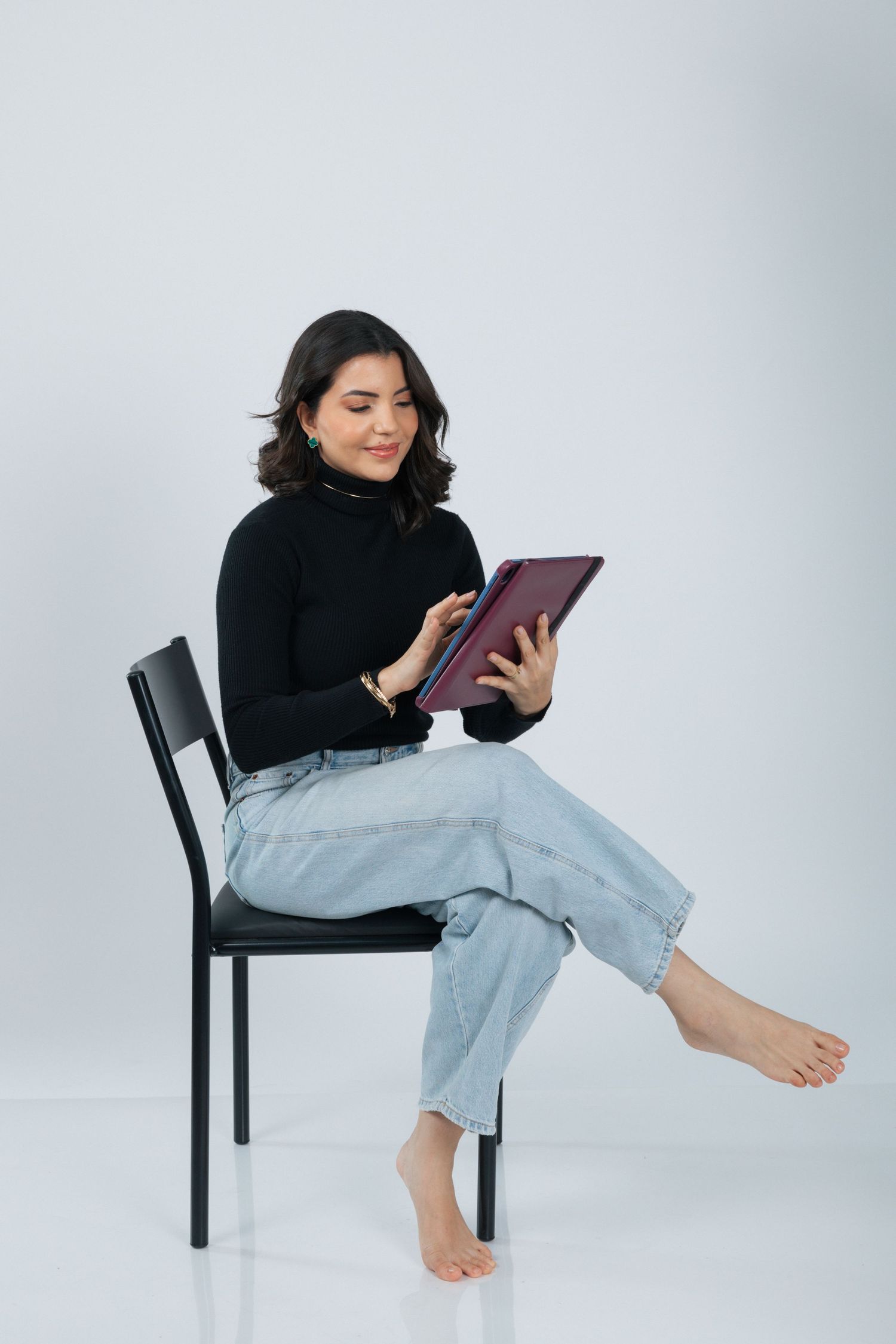 Woman in black turtleneck and jeans reading from a tablet while seated barefoot on a chair.