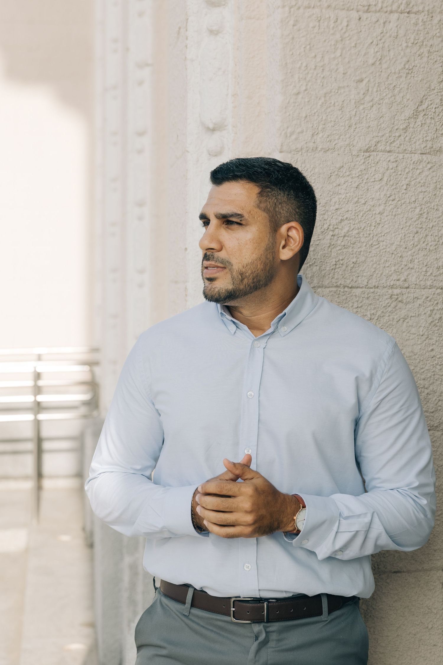 A person in a light blue dress shirt and gray slacks stands thoughtfully against a light-colored wall.