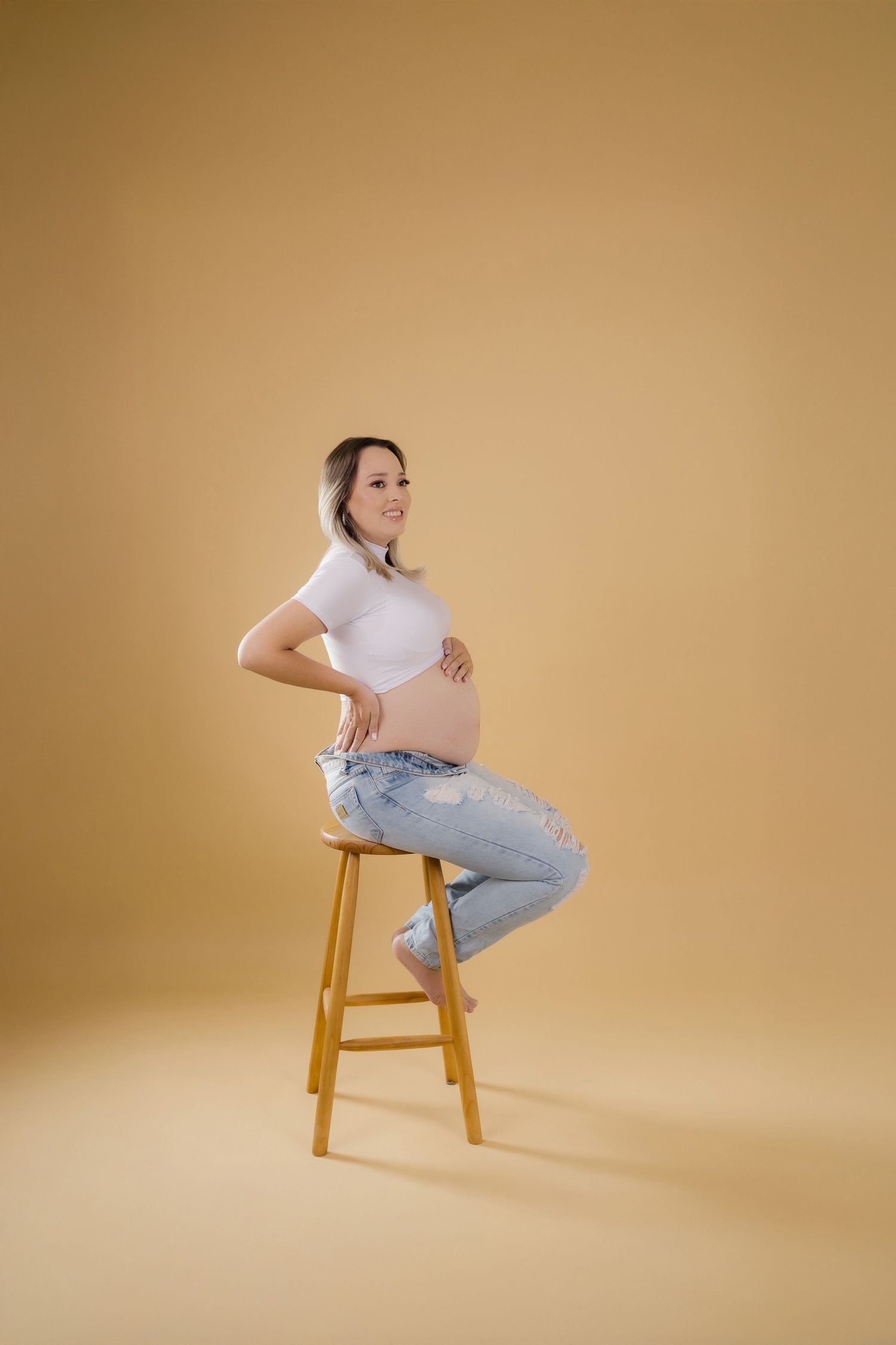 Woman in white crop top and jeans poses casually on wooden stool against warm brown studio backdrop.