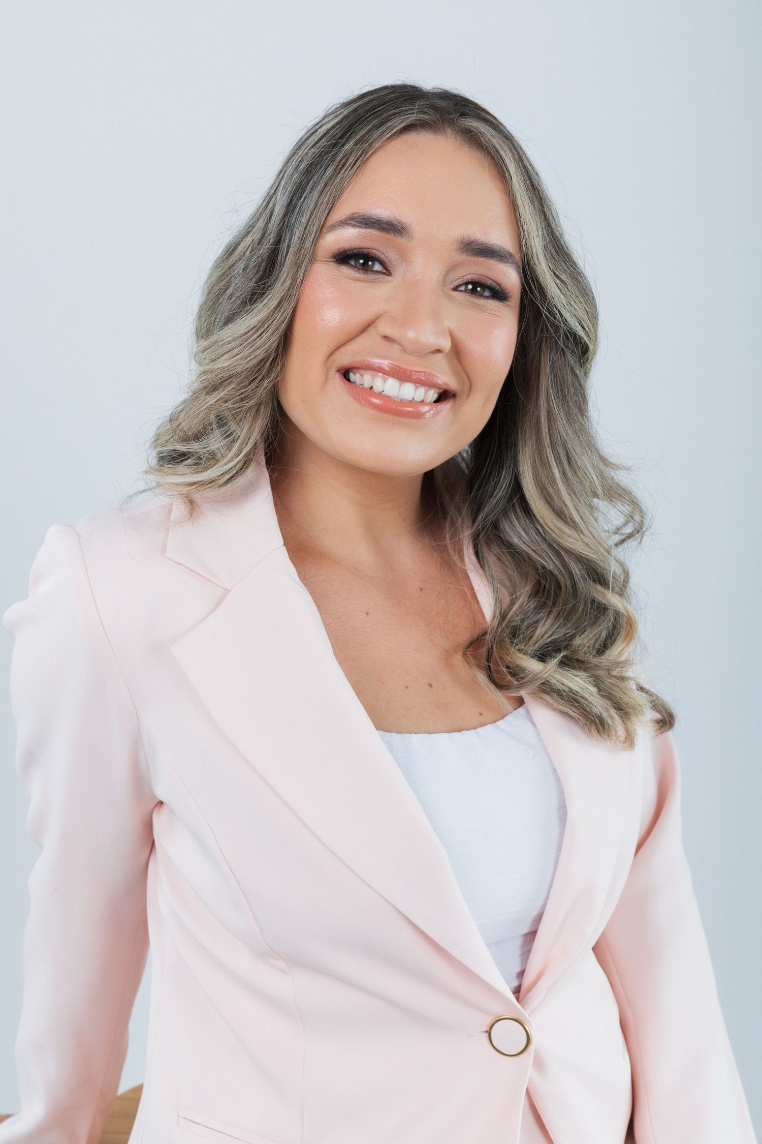Studio headshot of person with silver-toned wavy hair styled in loose curls wearing pink blazer and white blouse.