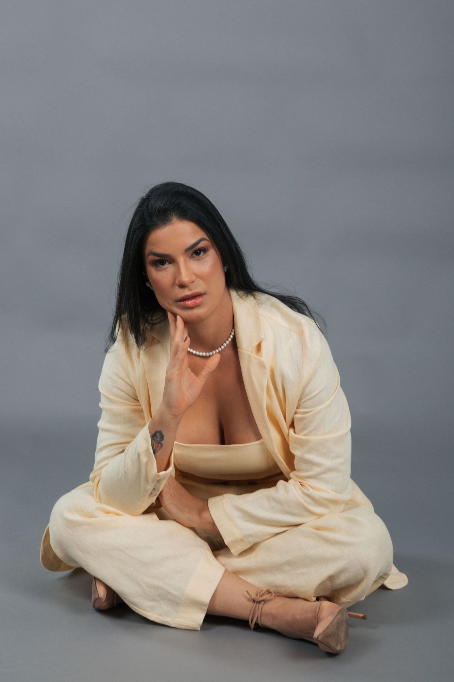 Person in cream-colored linen suit sits cross-legged on grey backdrop wearing pearl necklace and neutral toned top.