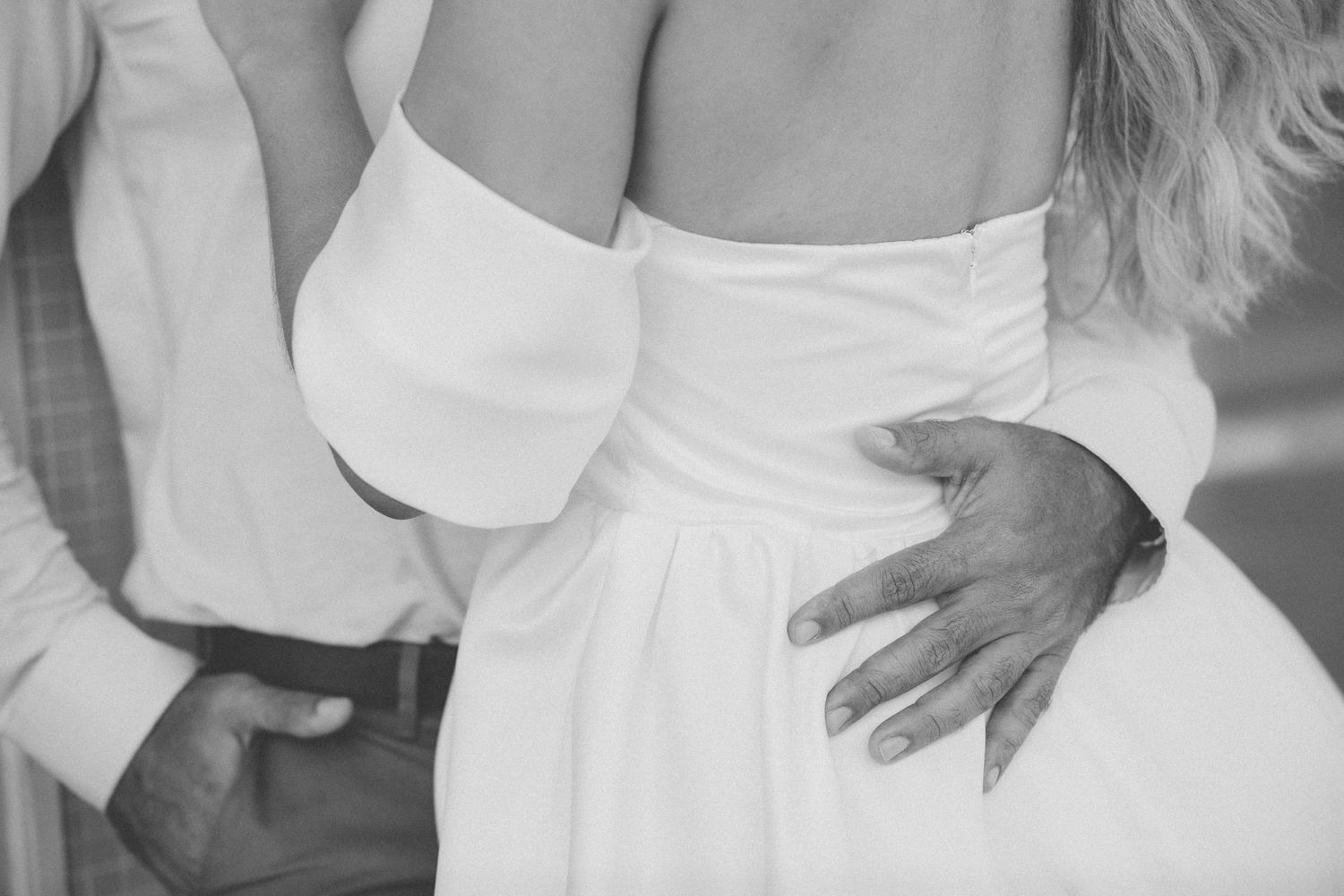 A black and white photograph showing hands tenderly embracing around a white dress during an intimate moment.