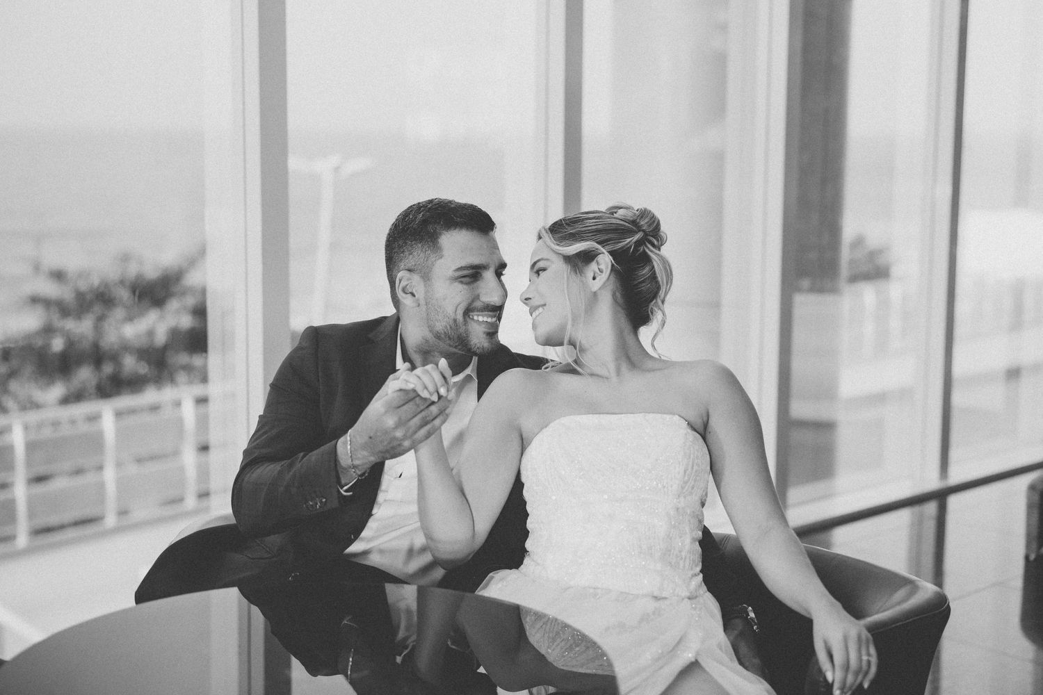 A couple shares a happy moment together in formal attire by large windows at an indoor venue in black and white.