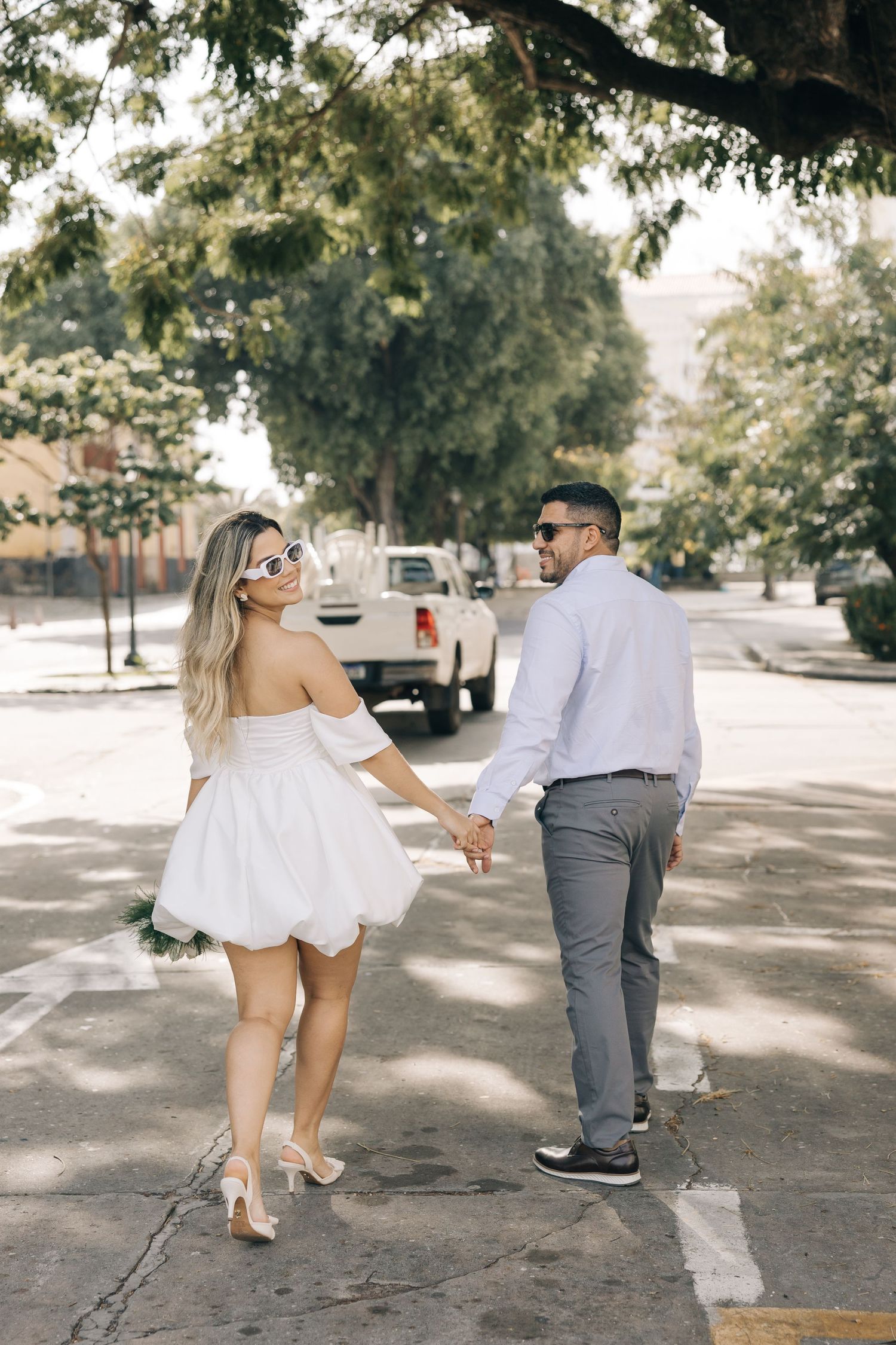 A couple holds hands while walking down a tree-lined street, with her in a white dress and him in grey pants.