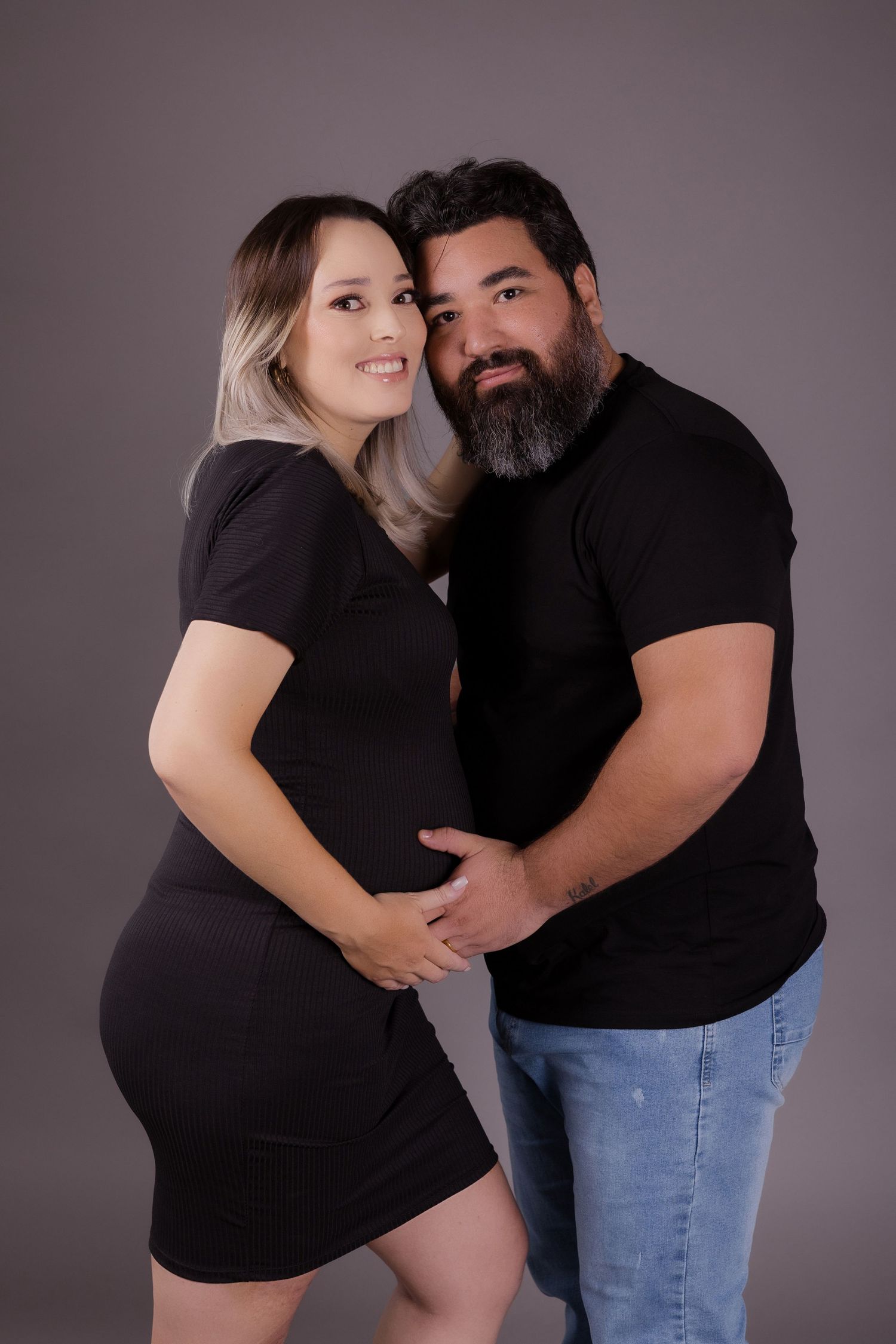 A couple in black outfits share an intimate maternity portrait against a dark background.