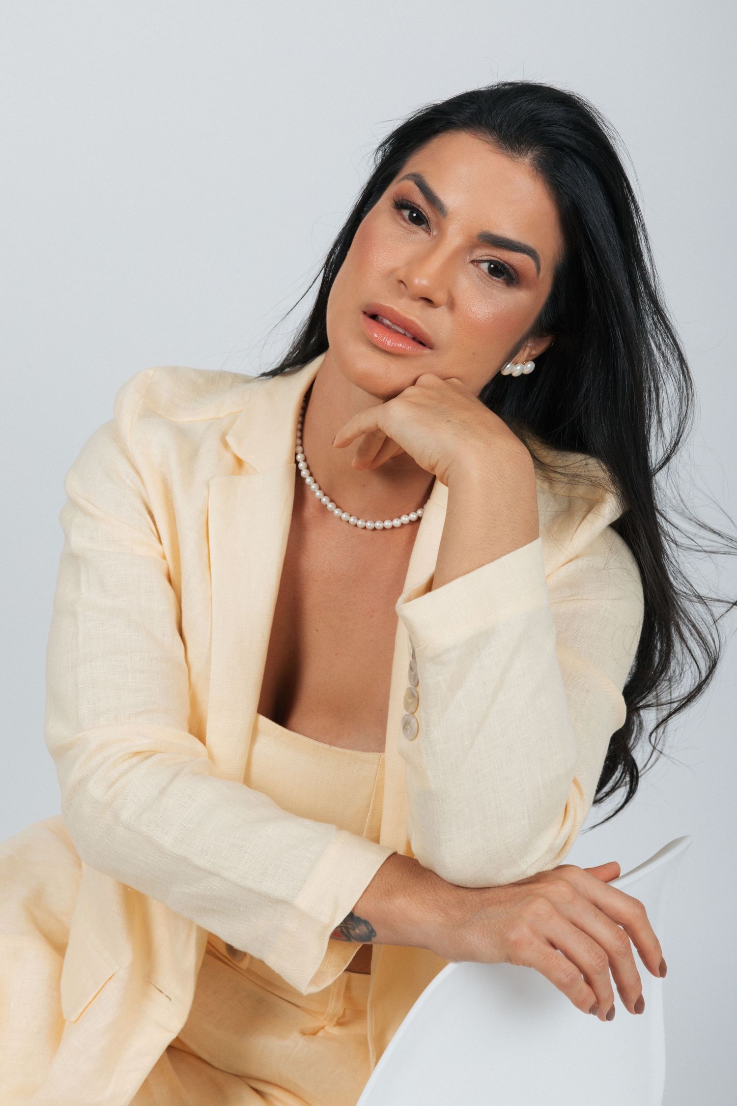 Model in cream colored suit and pearl necklace poses elegantly against studio background.