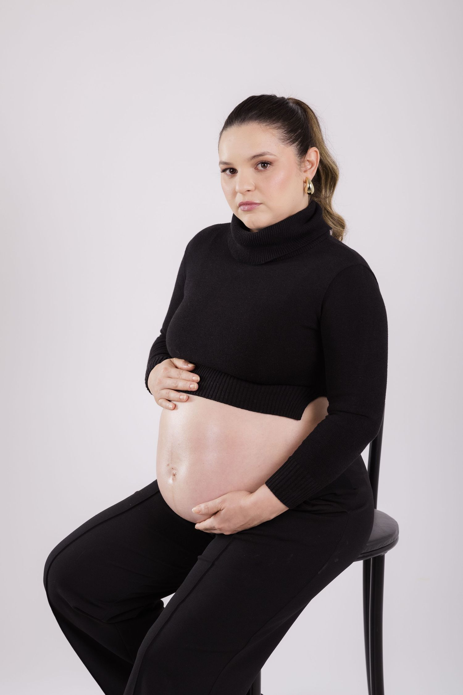 Pregnant woman in black turtleneck and pants sits on chair against light backdrop, cradling bare belly.