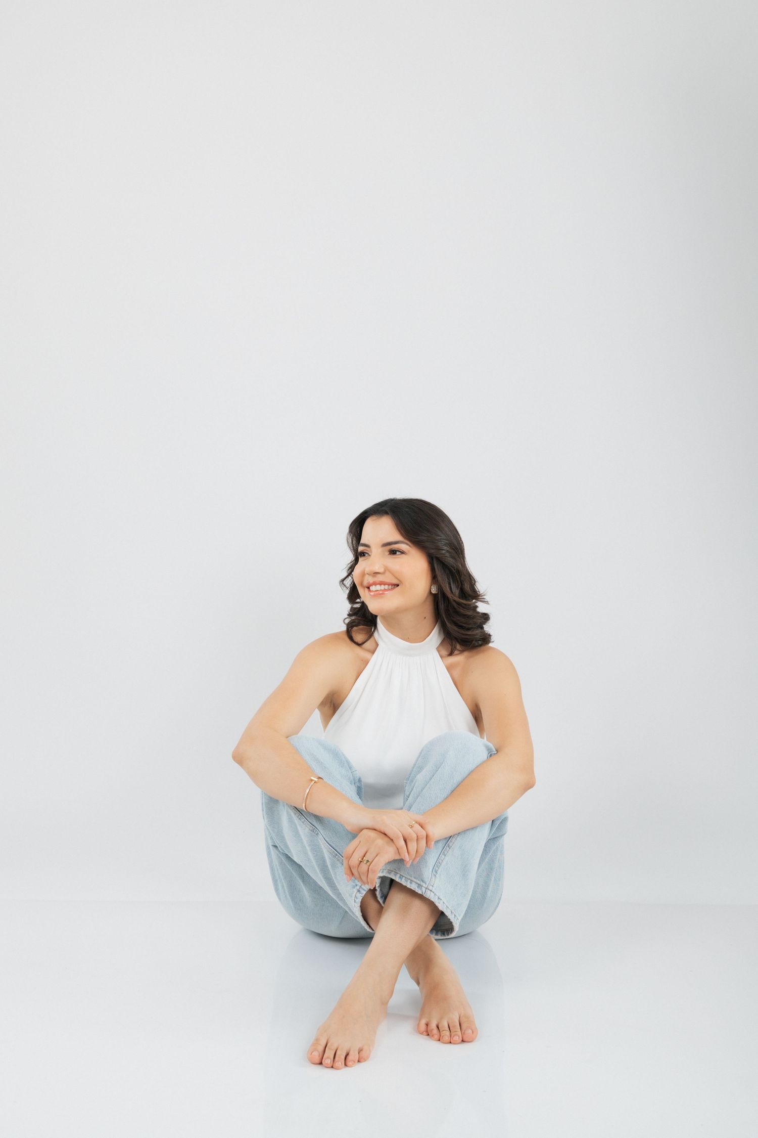 Woman in white halter top and light denim sits casually on the floor against white backdrop.