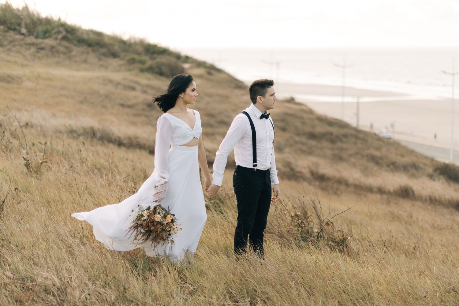 Wedding couple walks through coastal grassland with sea view and flowing white dress.