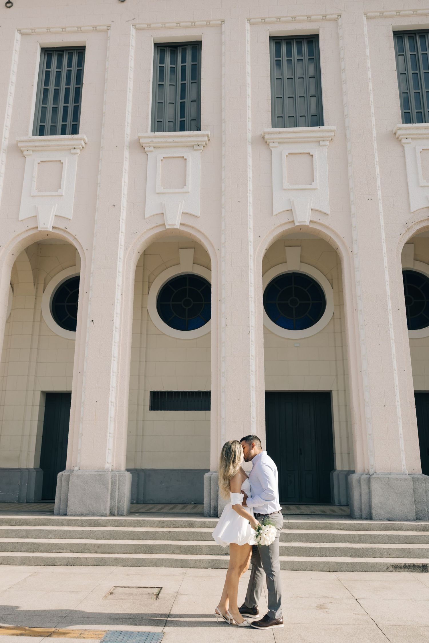 Couple embraces on steps of white classical building with arched windows and circular portals.