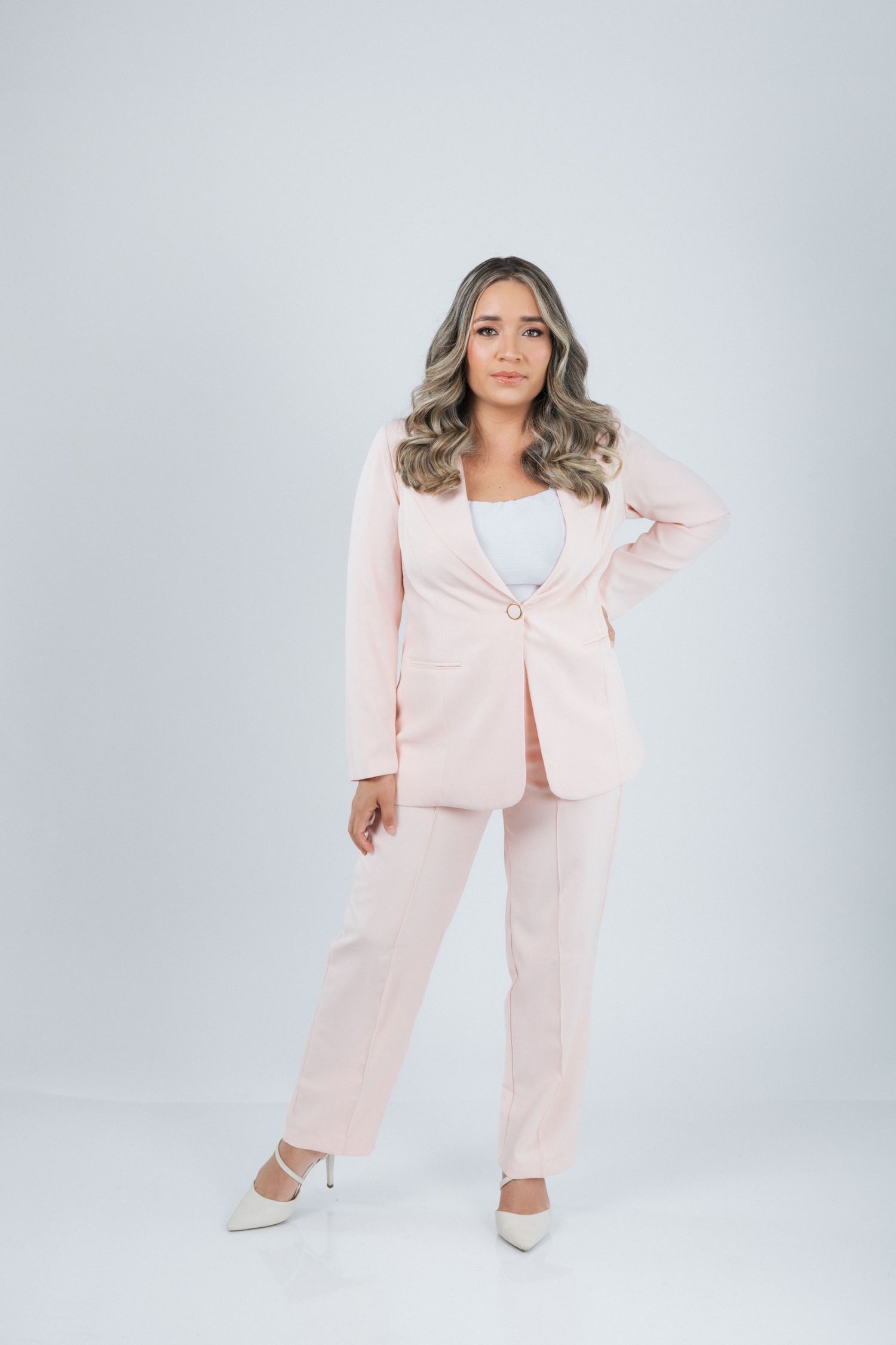 Professional fashion model wearing a pale pink business suit with white heels poses against light gray background.