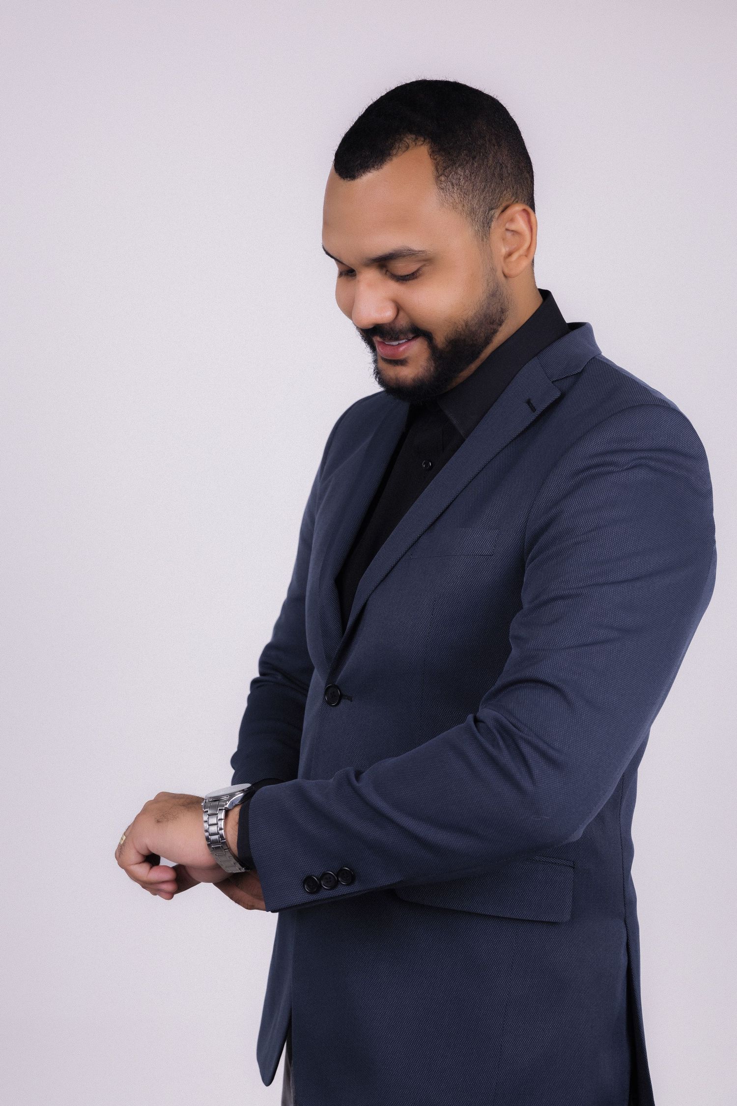 Professional wearing a navy blazer and black shirt checks wrist watch against light background.