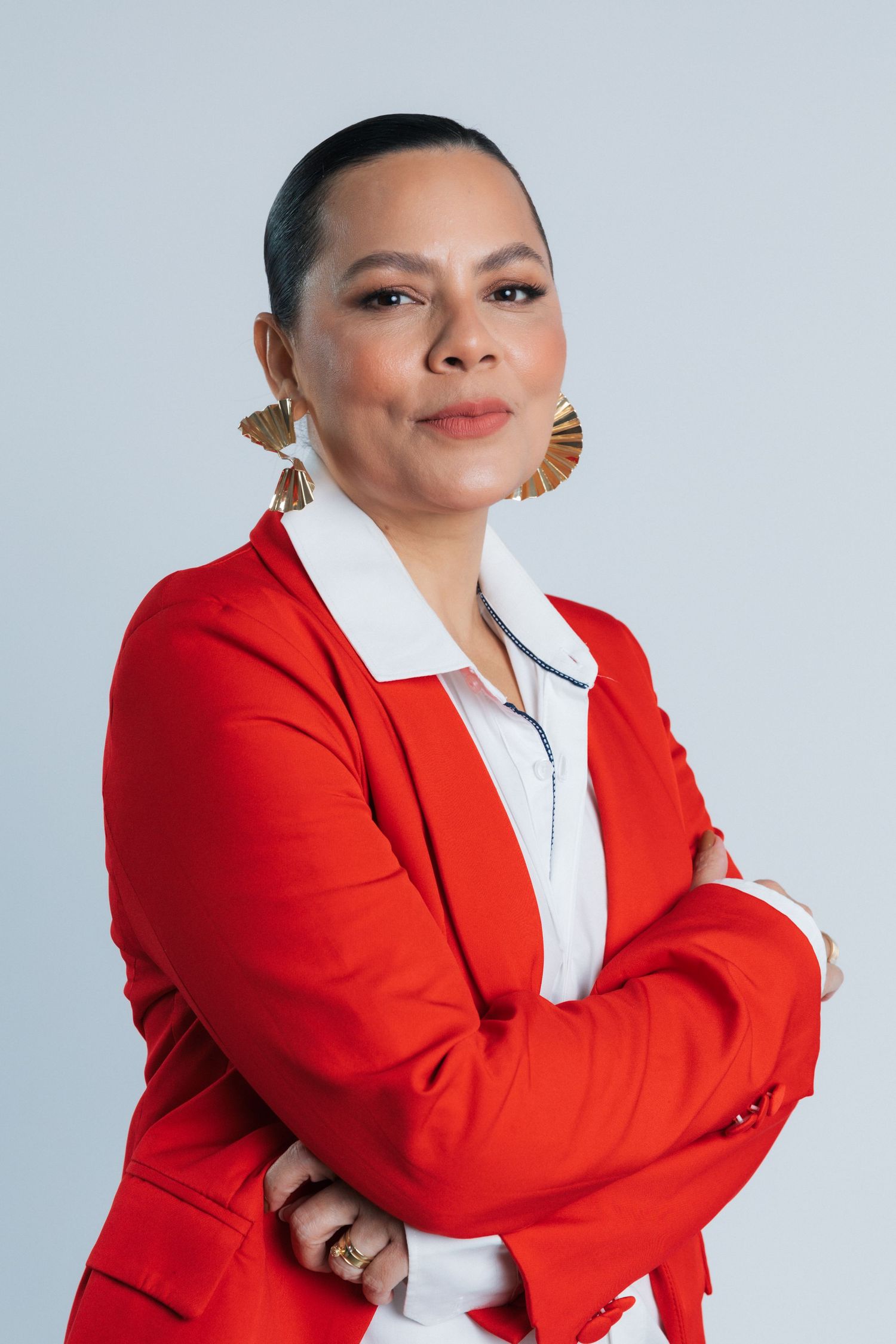 Confident pose in a vibrant red blazer with statement fan earrings against a gray backdrop.