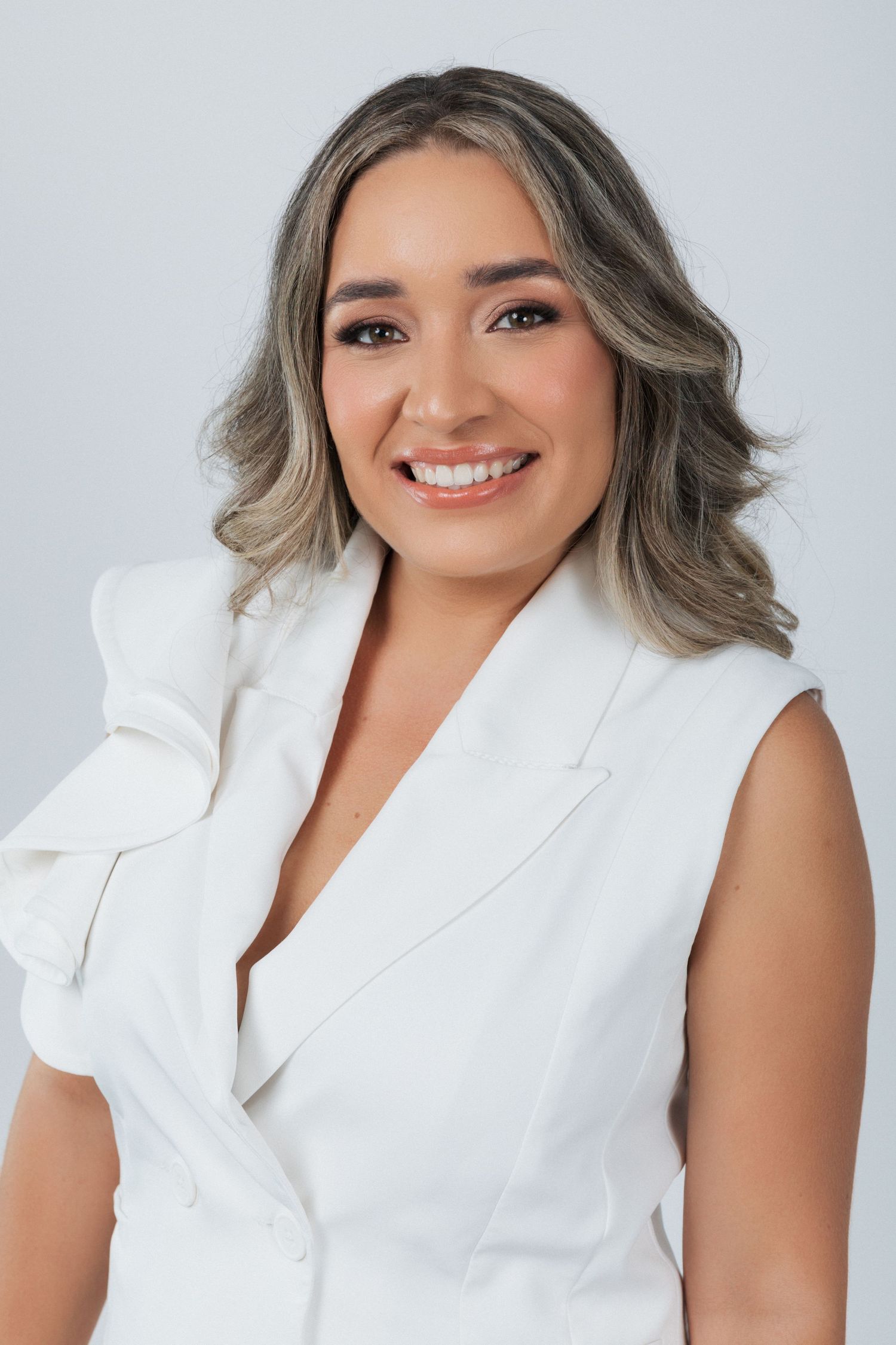 Professional headshot of a person wearing a white sleeveless blazer with gray wavy hair against a light background.