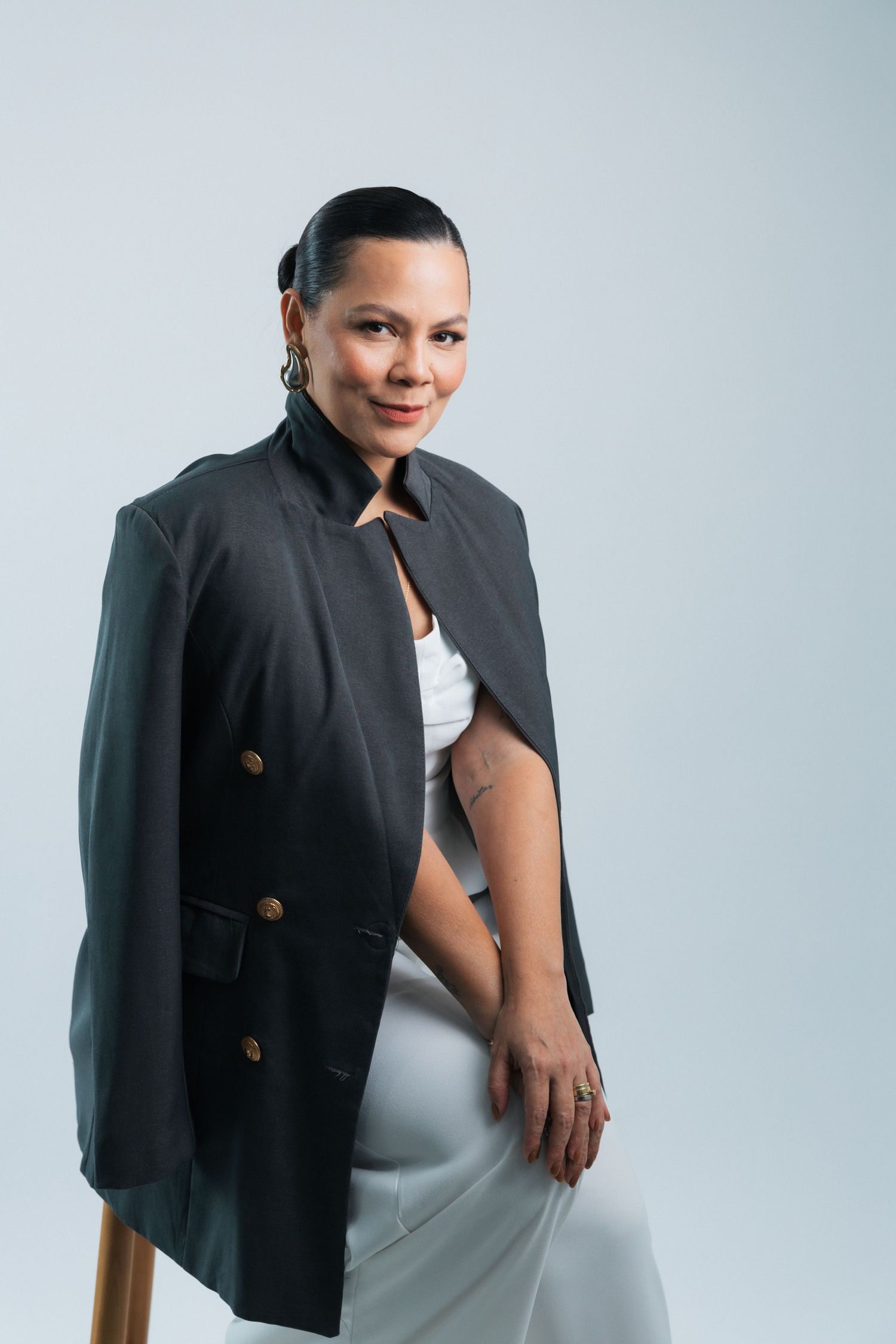 Professional woman in black blazer and white dress sits confidently on stool against light gray background.