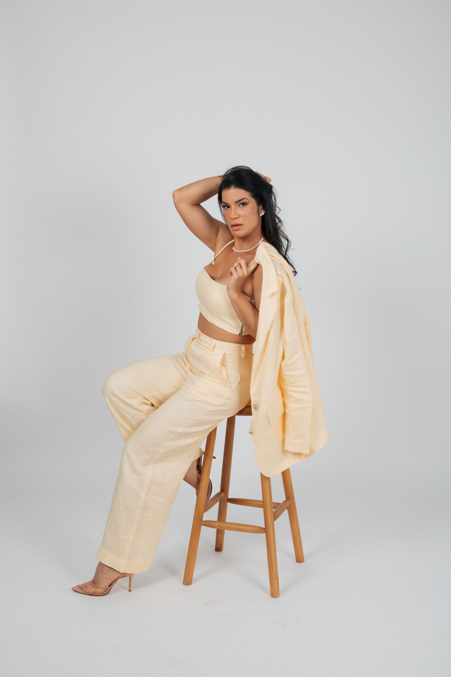 Model poses on wooden stool wearing cream-colored two-piece outfit with matching blazer against grey backdrop.