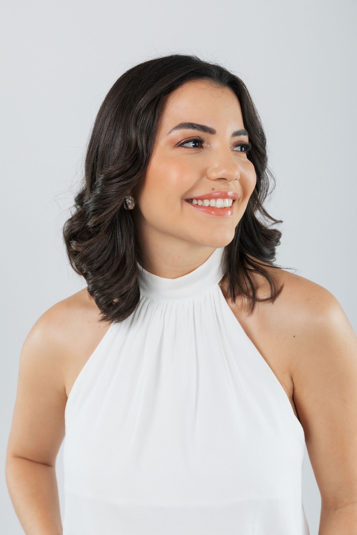 Portrait of a model with shoulder-length dark wavy hair wearing a white halter top against a light gray backdrop.