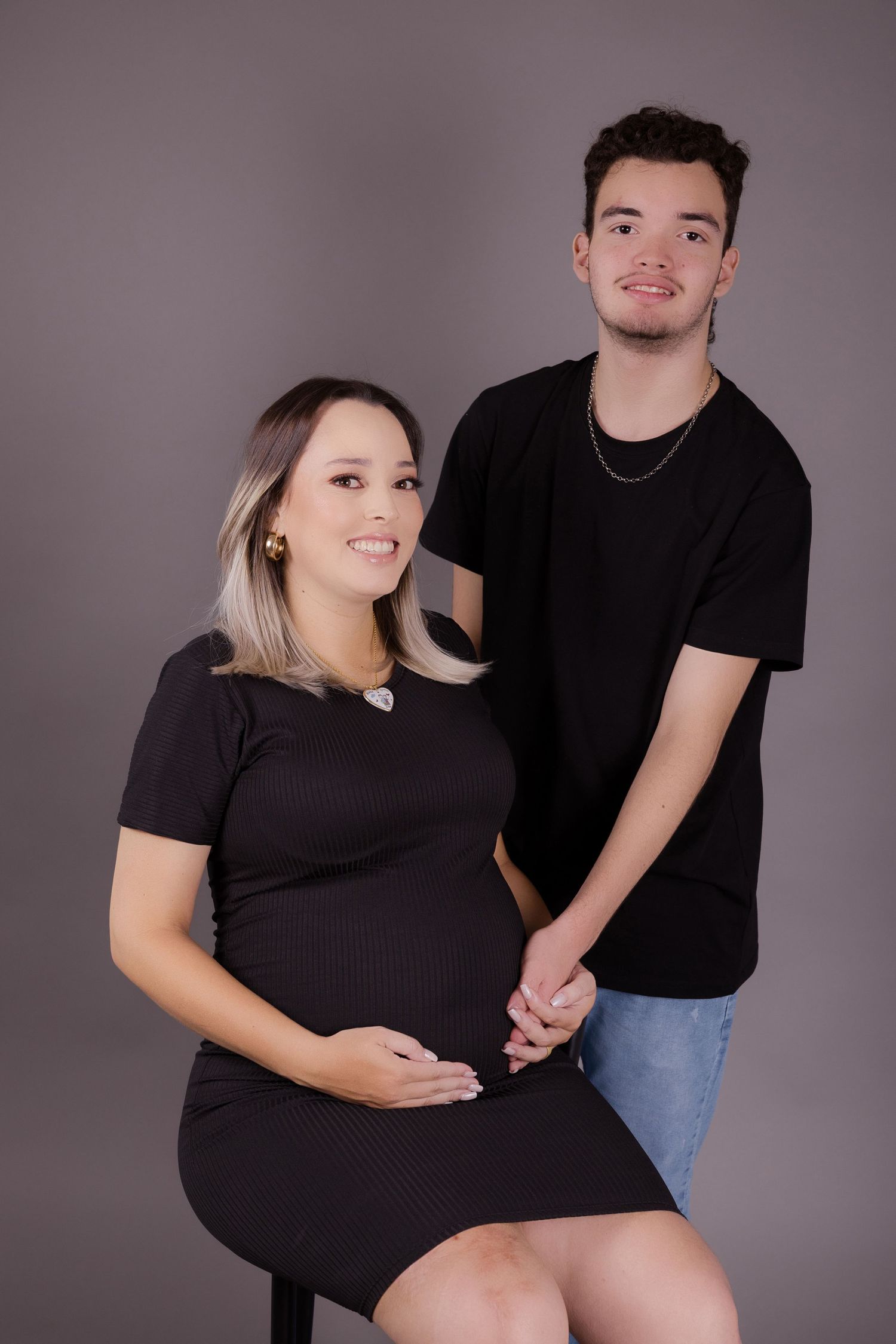 A pregnant woman in a black dress poses with a partner wearing dark clothes during a maternity photoshoot.
