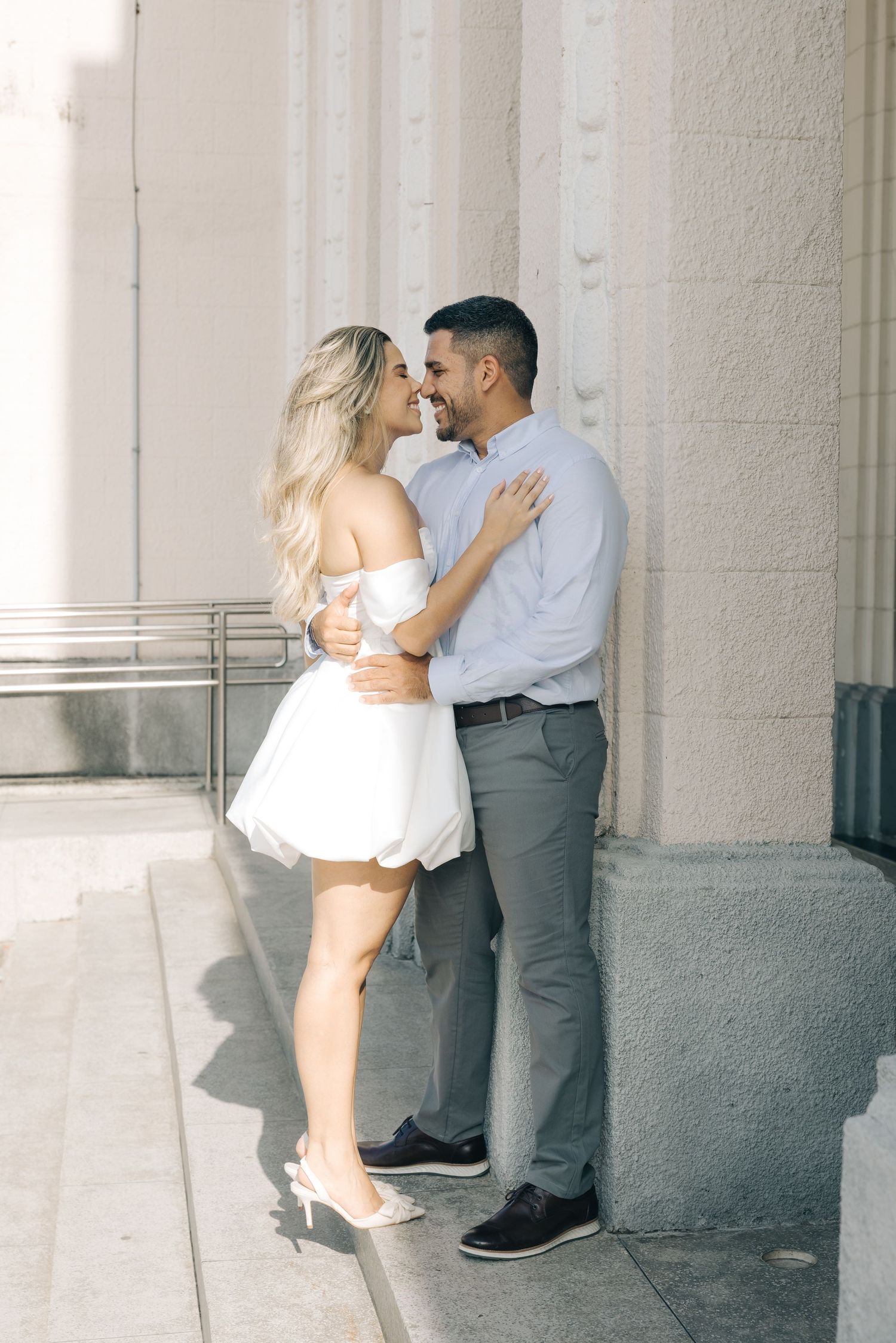 A romantic couple dressed in white and grey share a tender moment against a white stone building wall.