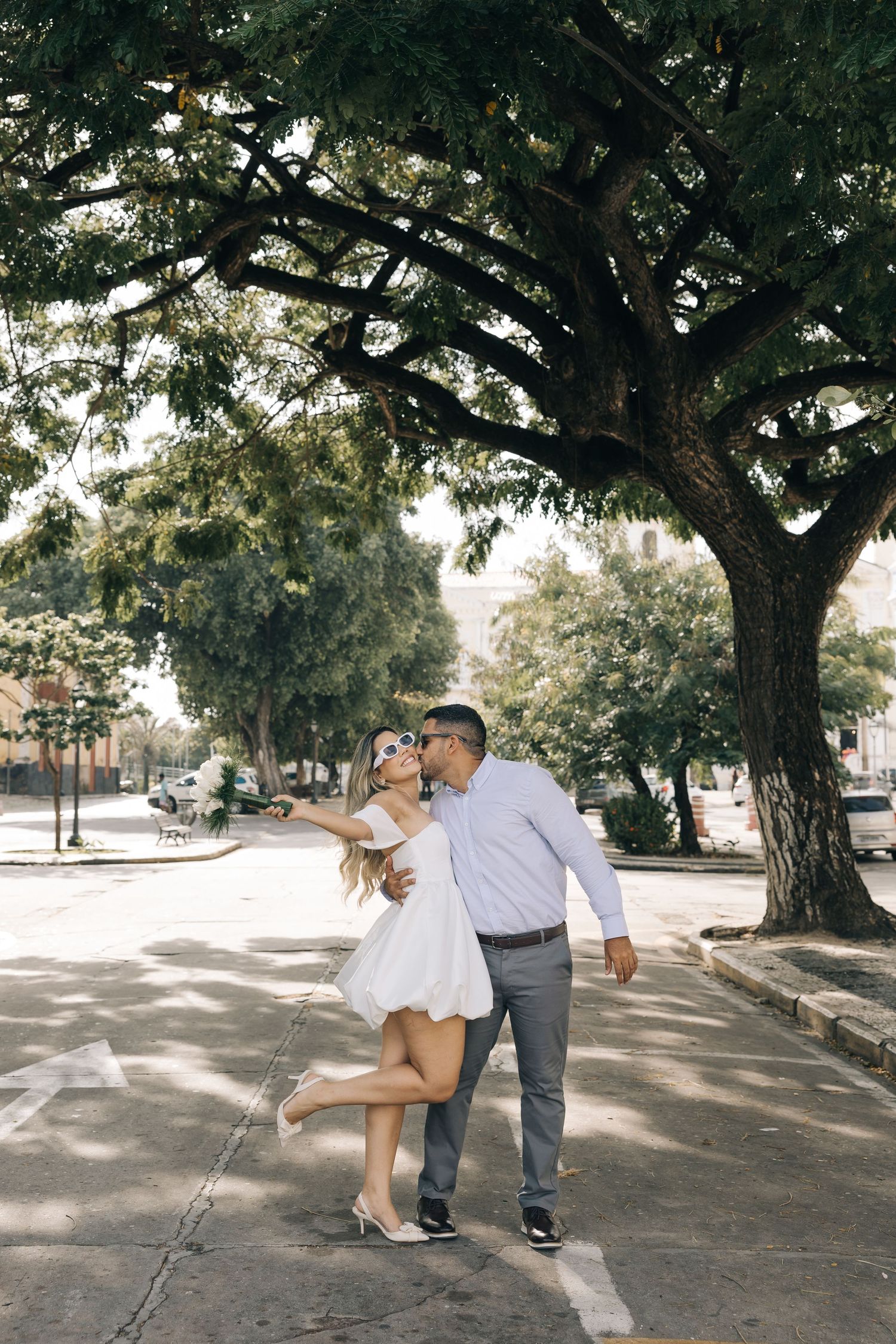 A couple shares a playful moment together on a tree-lined street on a sunny afternoon.