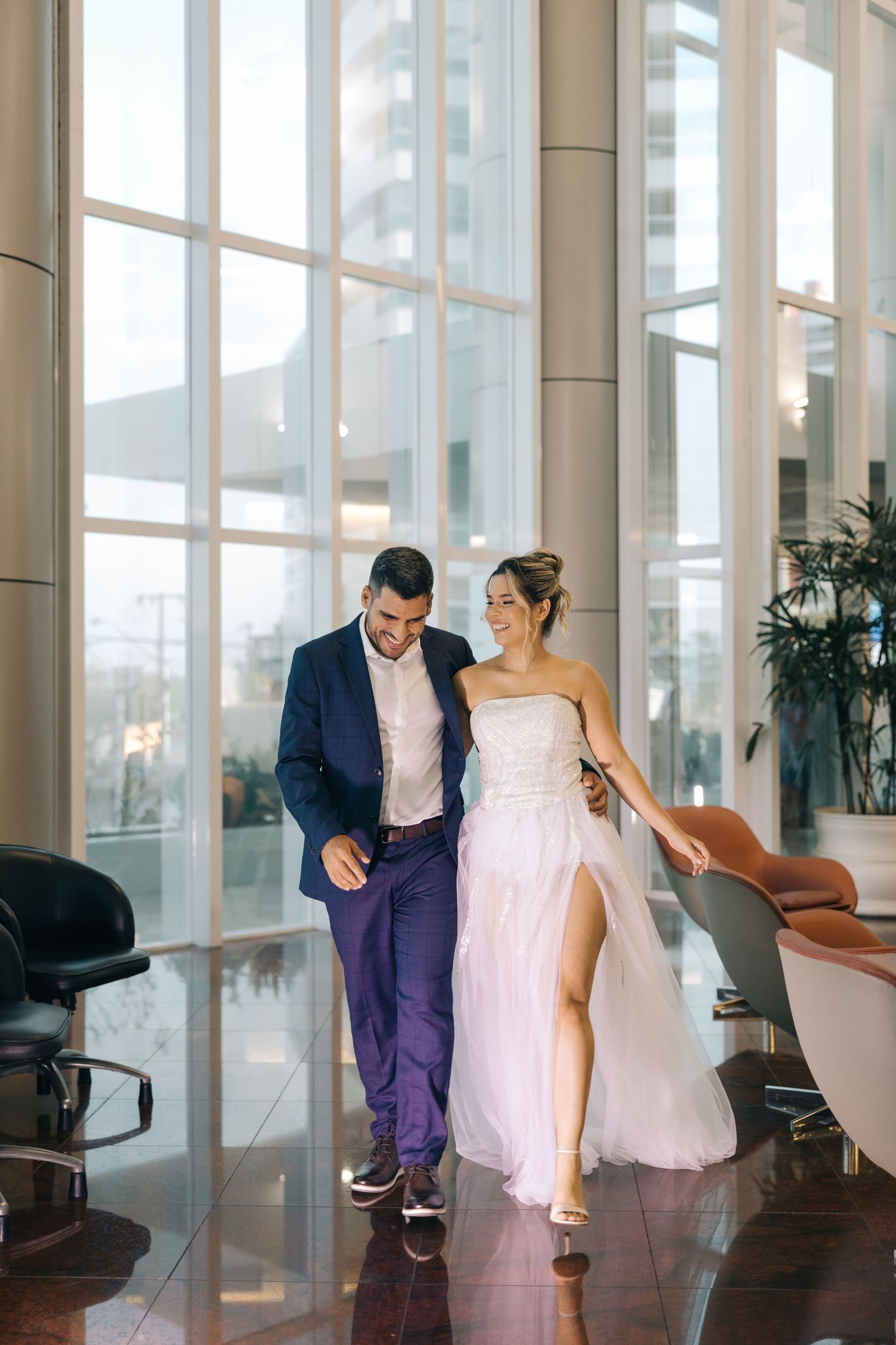 A couple in formal attire walks through an elegant modern lobby with floor-to-ceiling windows.