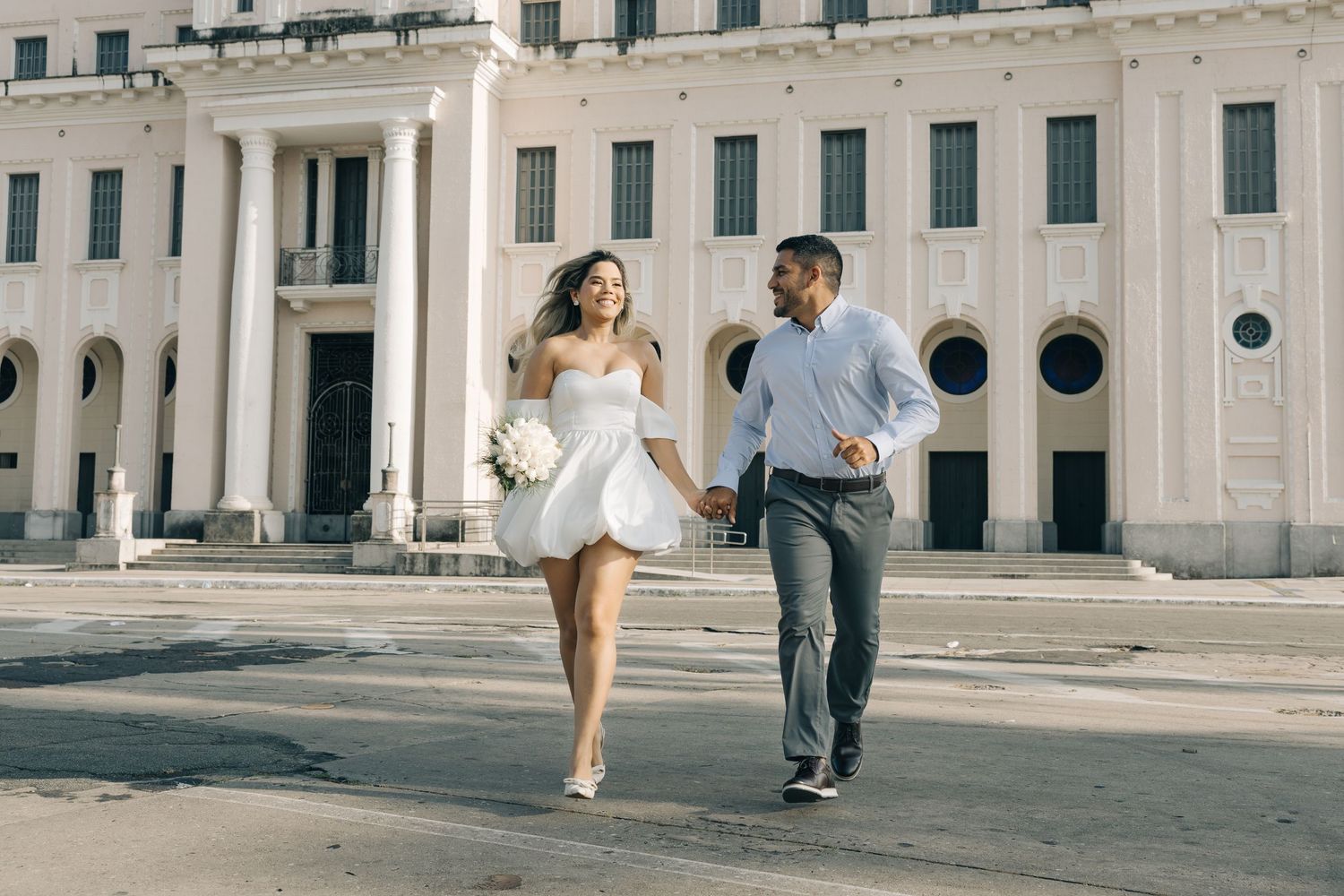 A couple runs playfully together outside a grand white building with classical architecture.