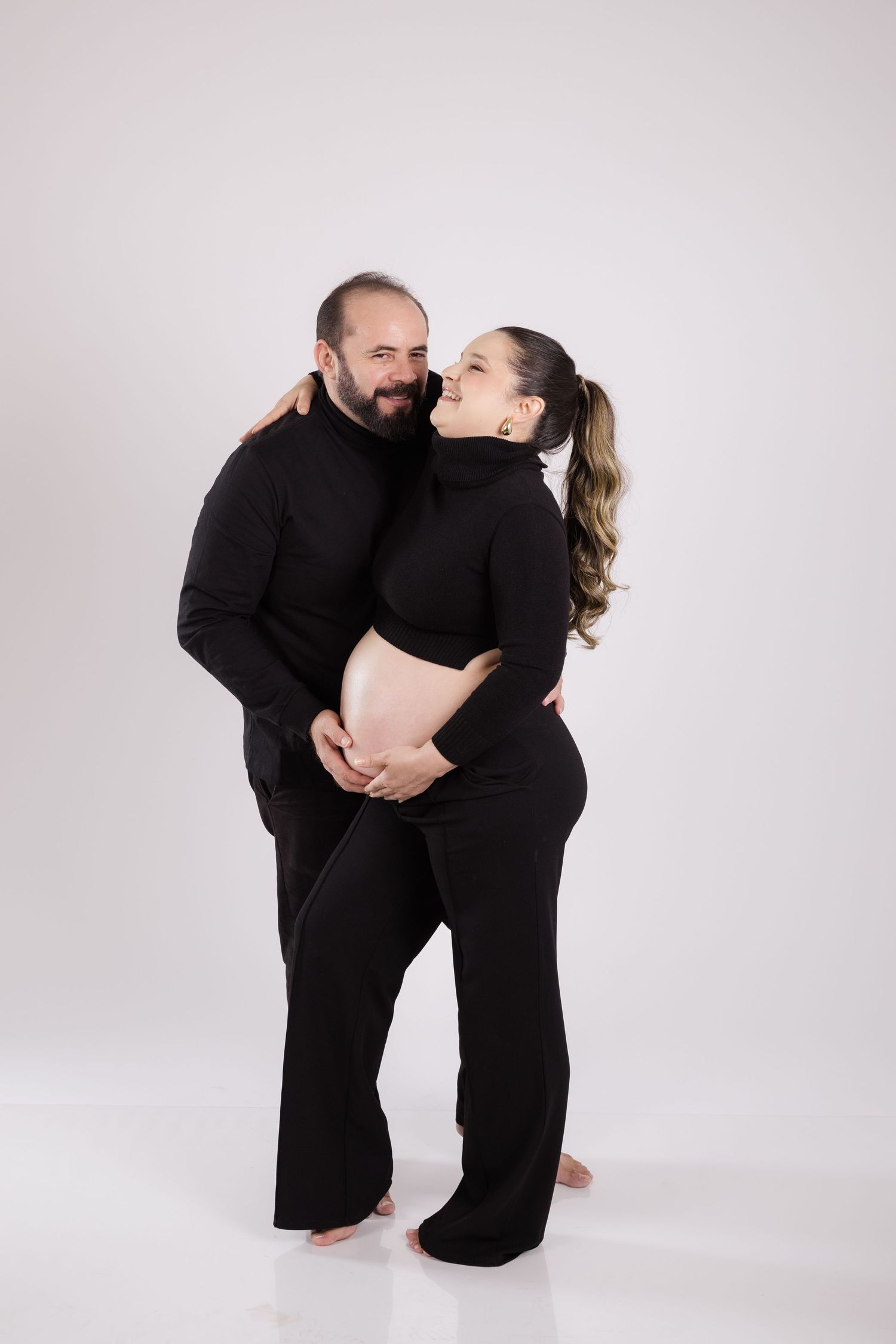 A couple wearing matching black outfits embrace while standing in a bright studio setting.