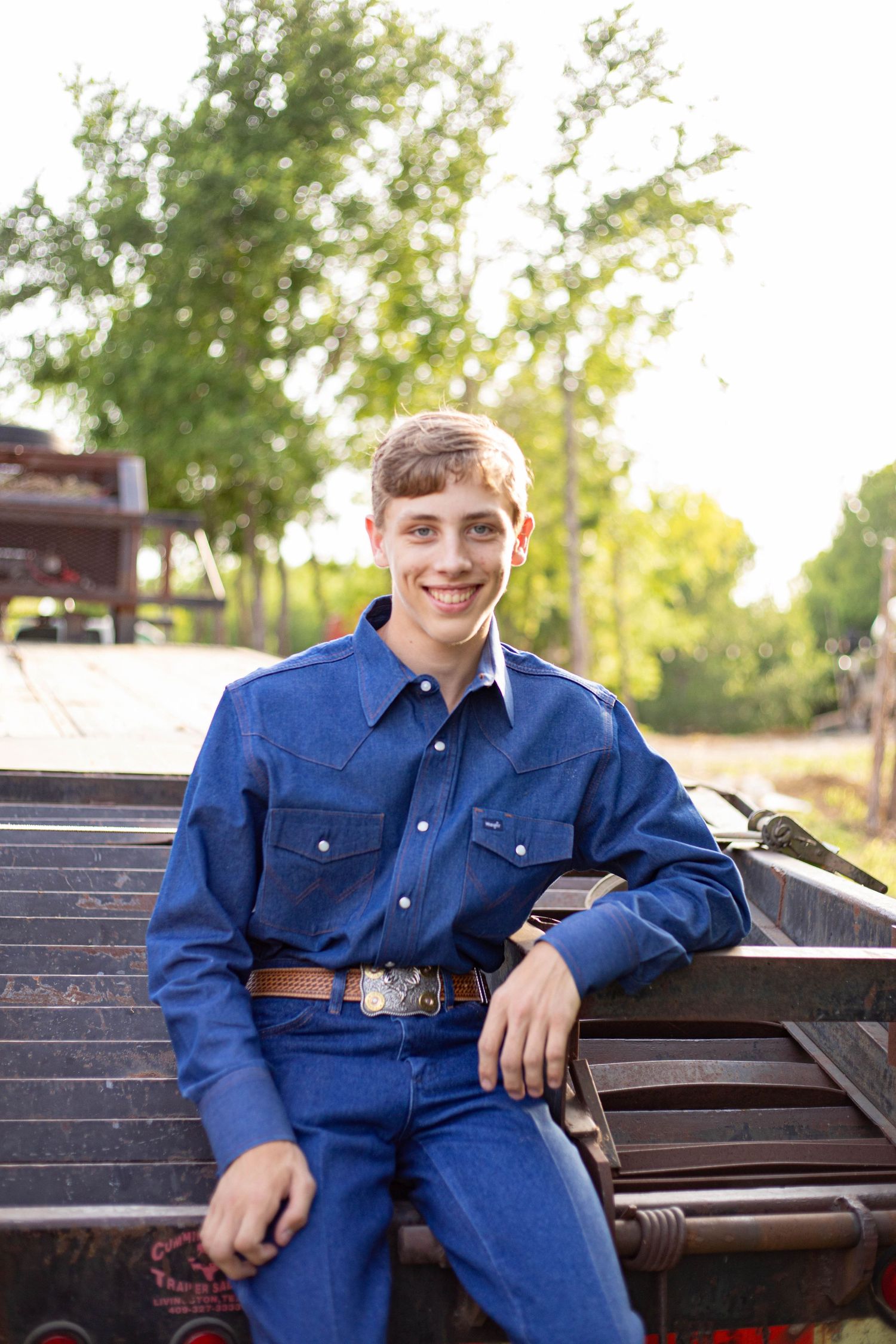 Smiling teenage boy in blue denim outfit with western belt sitting on a wooden bench outdoors.
