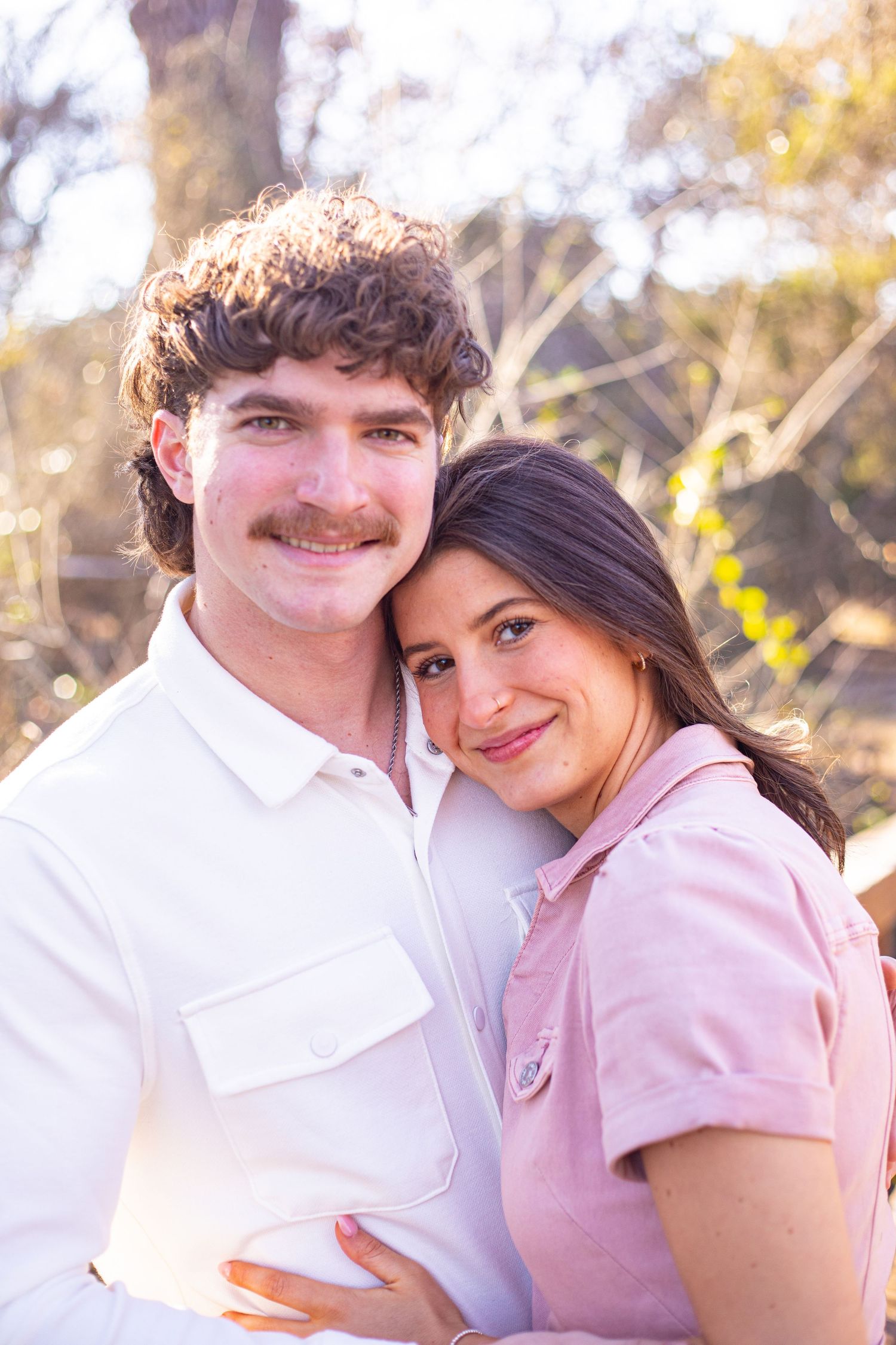 A couple in pink and white outfits share an intimate moment in golden sunlight.