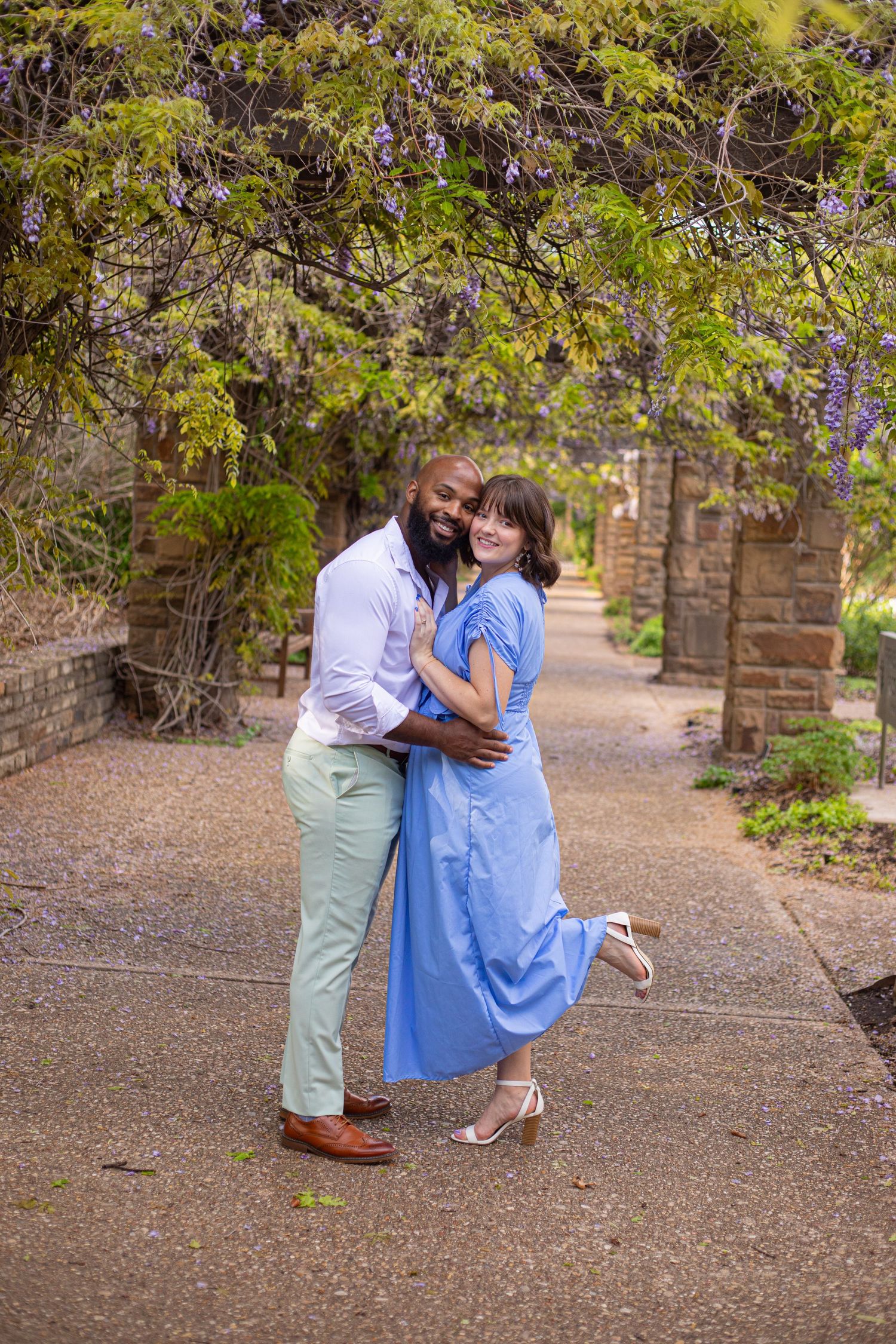 A couple embraces on a garden path with blooming wisteria and stone walls in the background.
