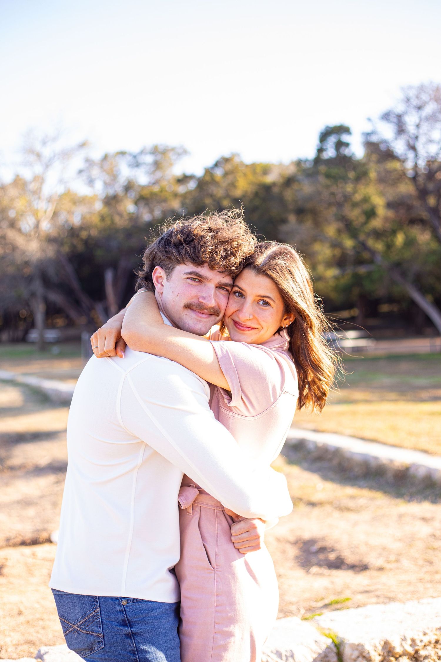 A couple shares a tender moment while wearing white and pink outfits in a natural outdoor setting.