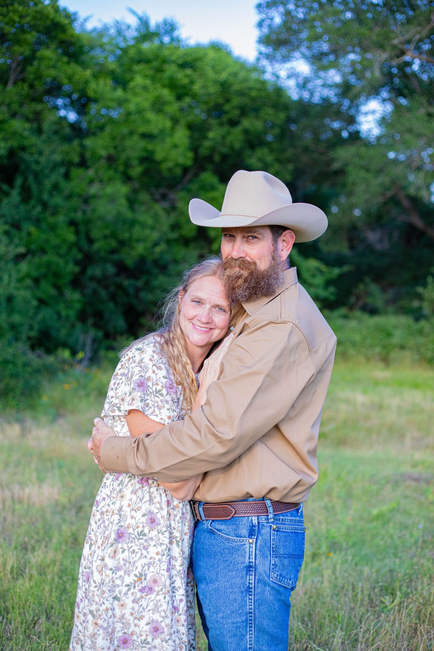 A bearded man in a cowboy hat and woman in a floral dress embrace outdoors in a green field.