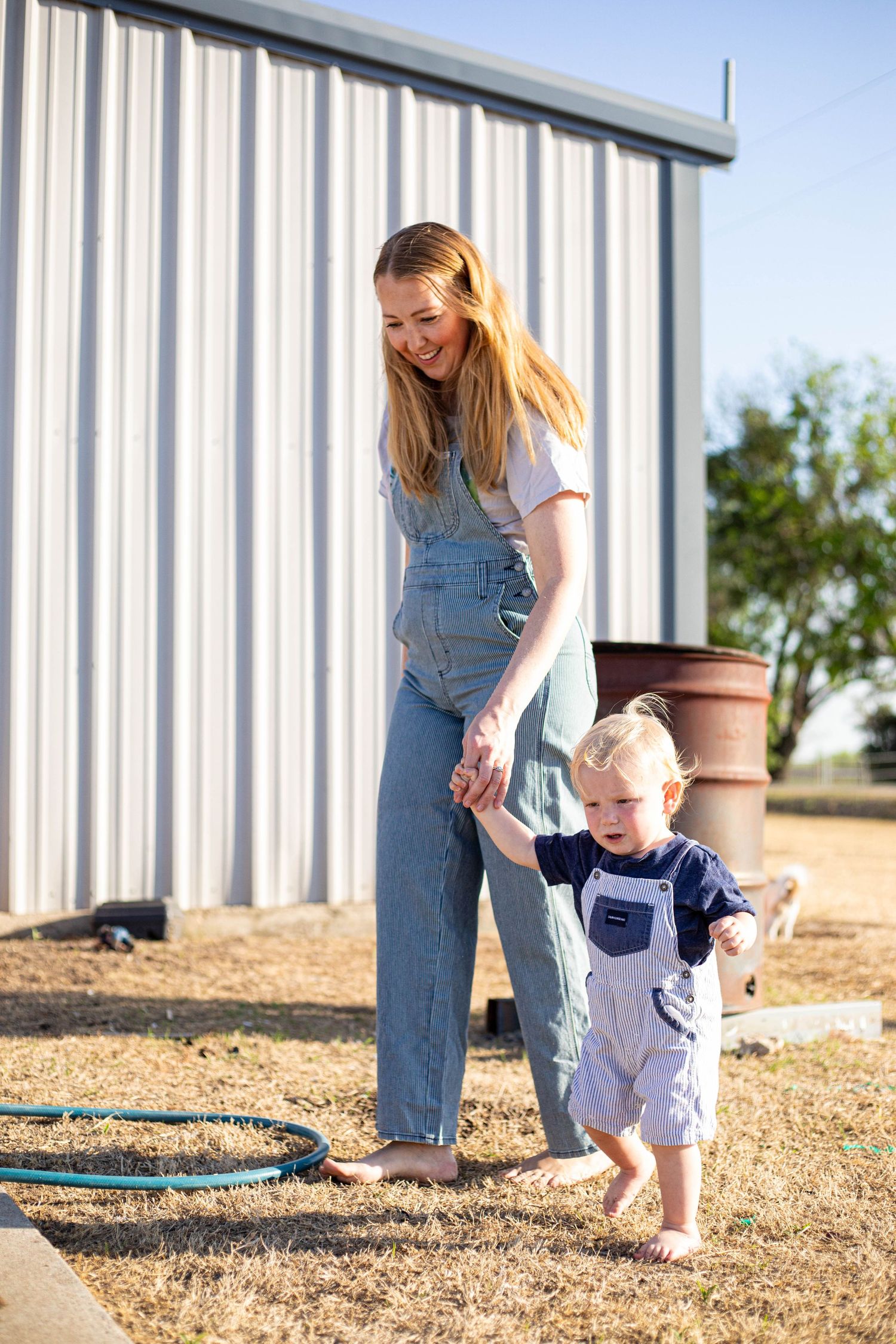 A smiling woman in denim overalls helps a toddler take first steps outdoors near a metal shed on a sunny day.