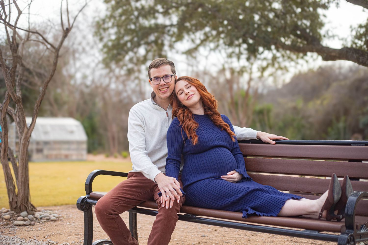 Pregnant couple sitting on a park bench, woman in blue maternity dress leaning on man in white shirt.