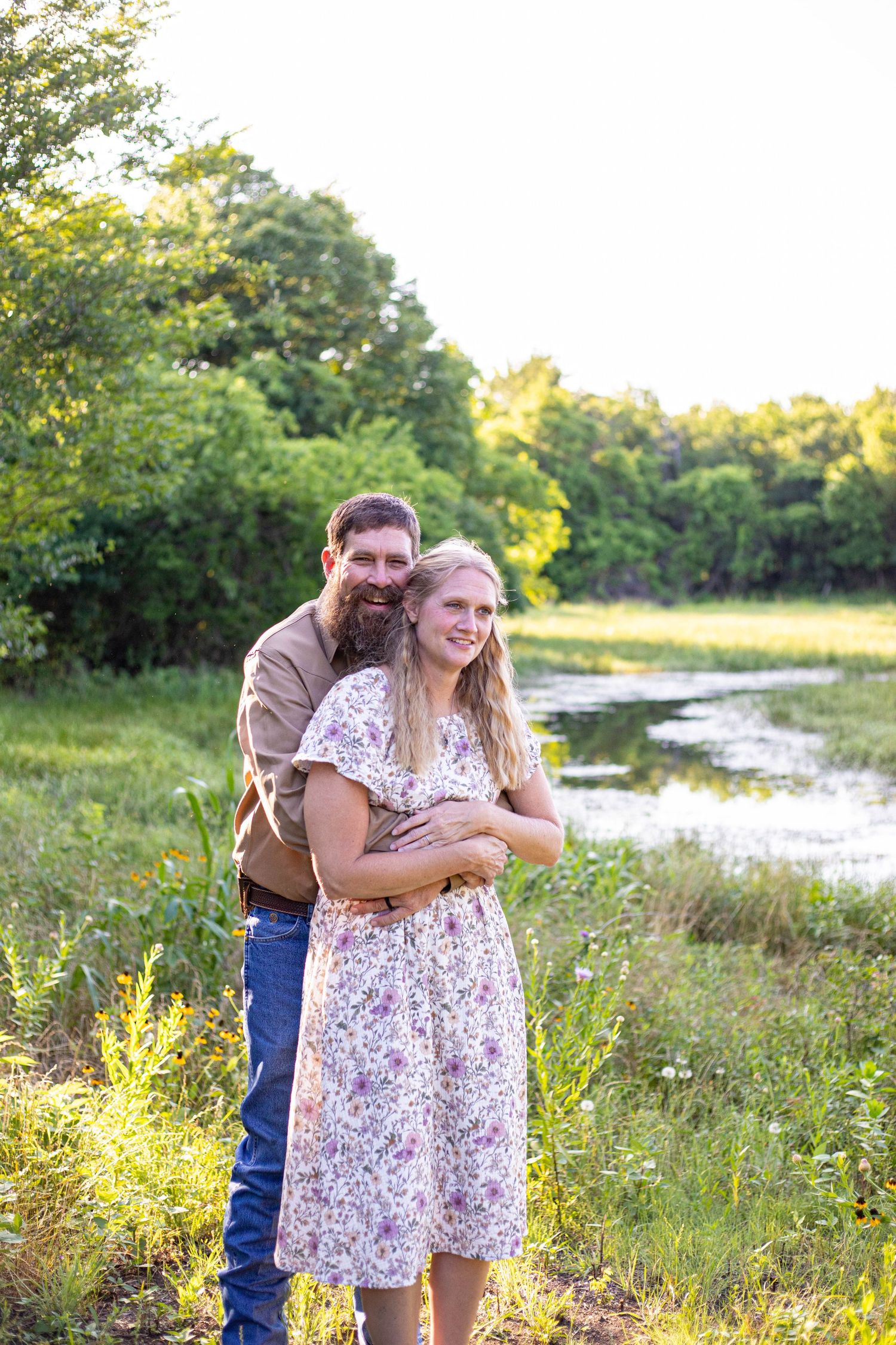 Couple embracing outdoors near a creek, woman in floral dress, man in brown shirt and jeans, lush green trees background.