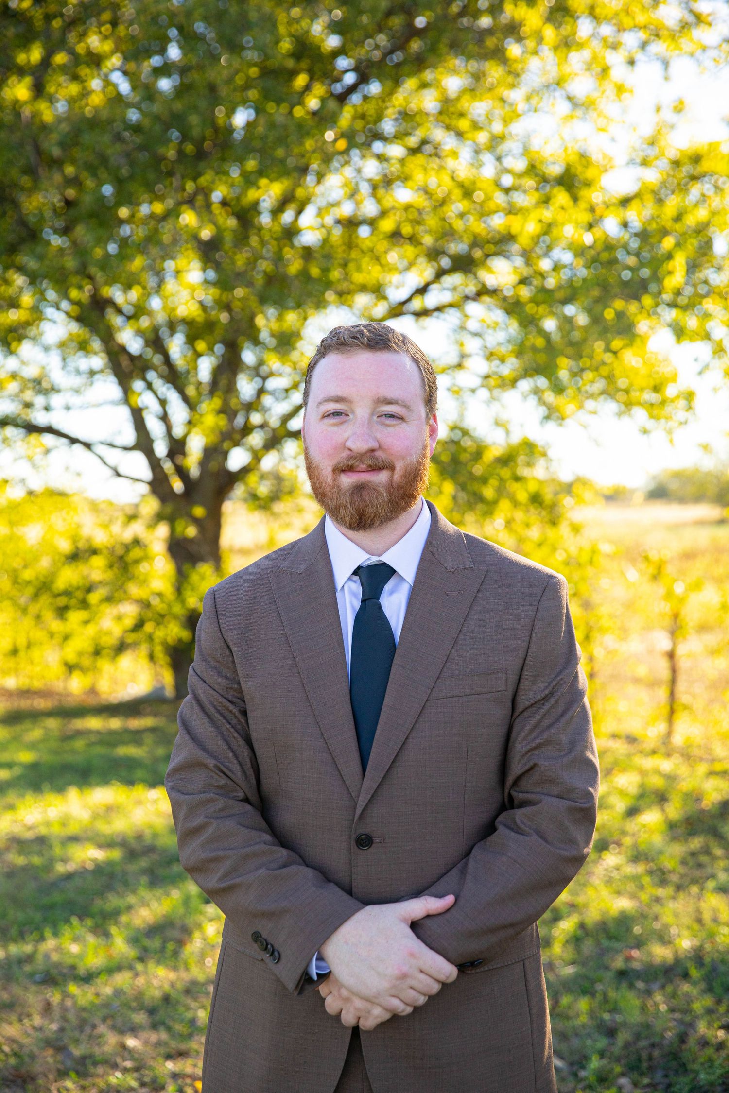A bearded man in a brown suit and dark tie smiles outdoors with green trees in the background.