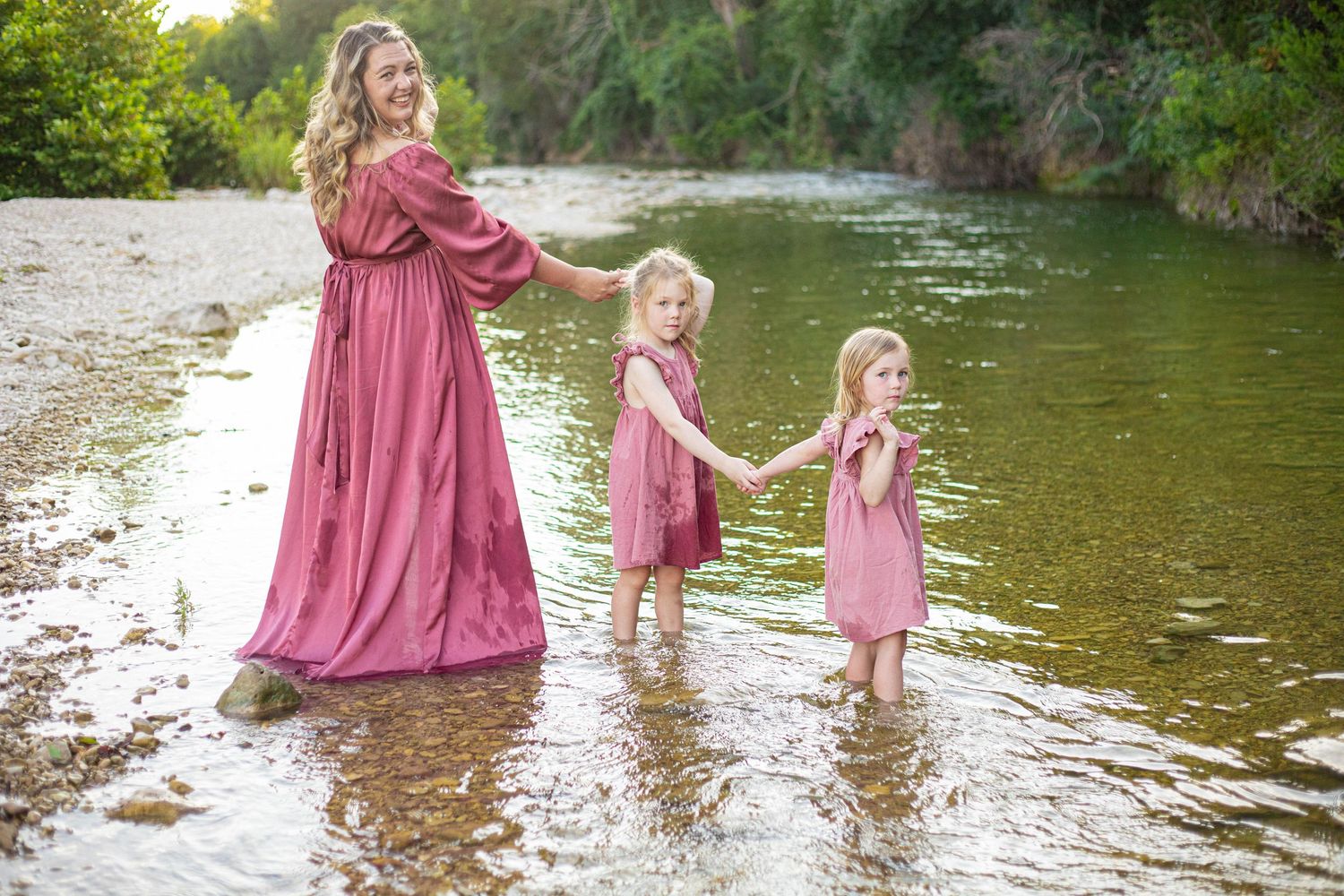 Family members in flowing pink dresses walk along the peaceful riverbank at sunset.
