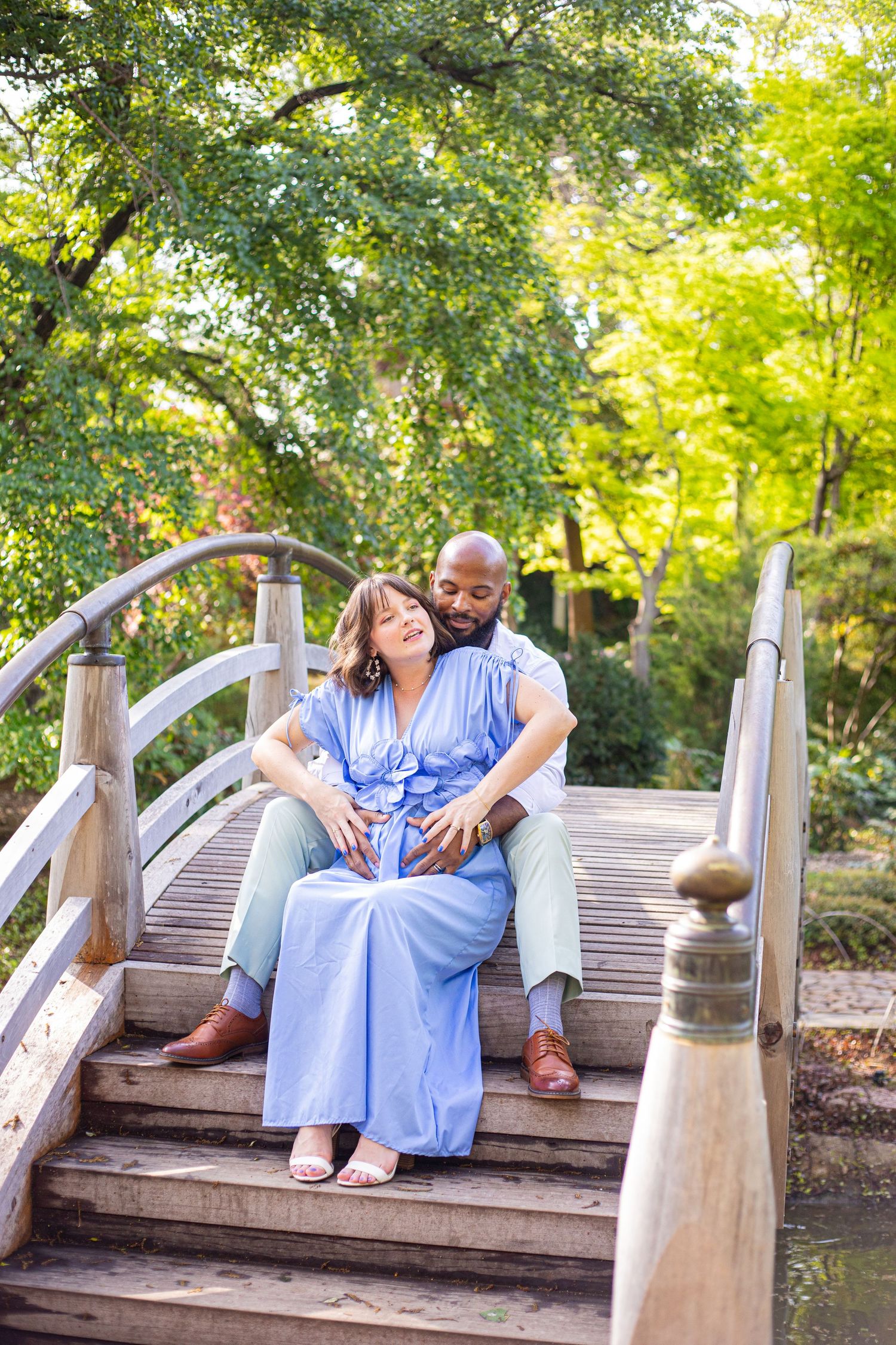 Couple cuddling on wooden bridge surrounded by green trees in garden setting.