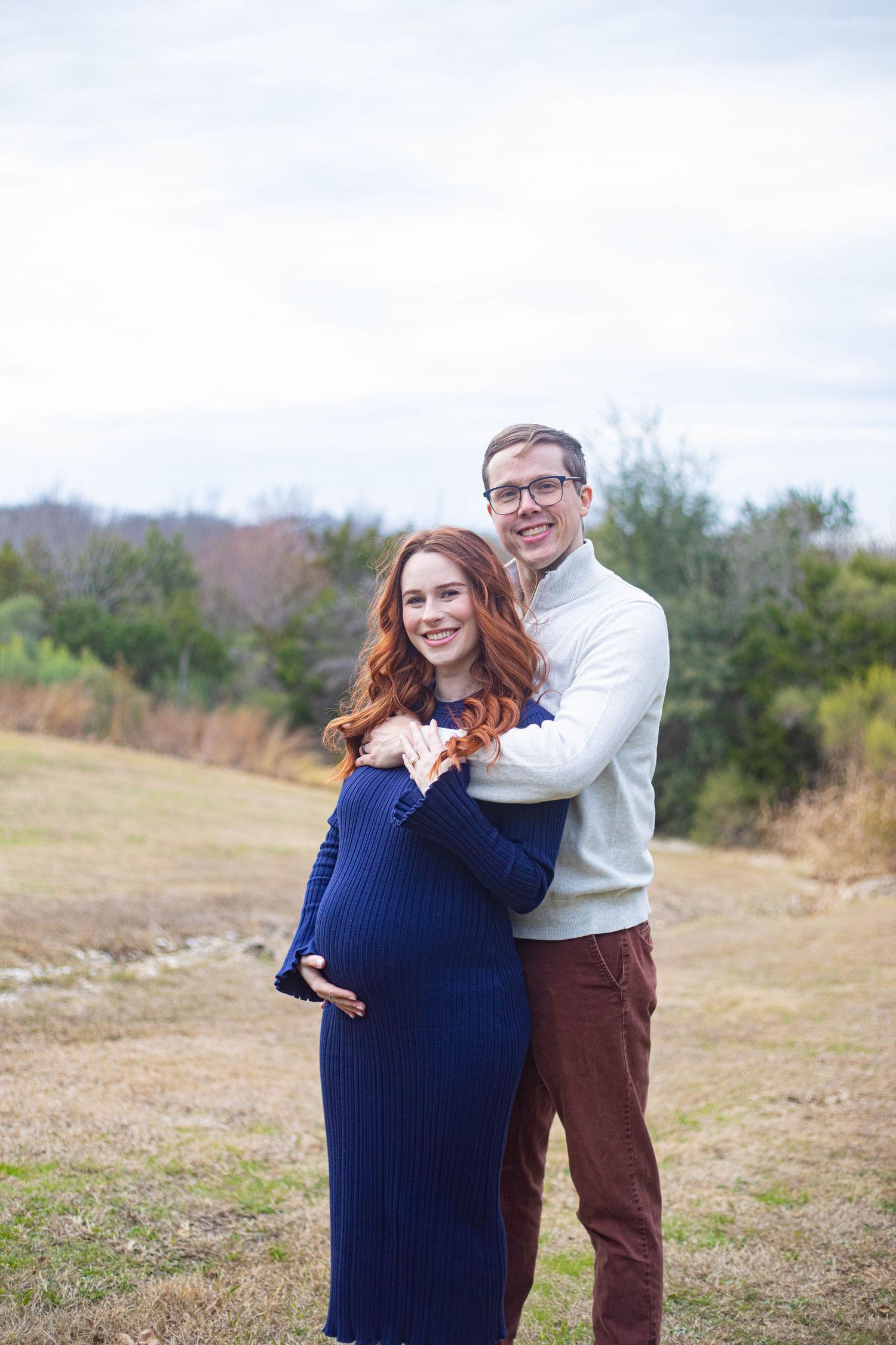 Smiling pregnant woman in navy dress with partner embracing outdoors in a grassy field during maternity photo session.