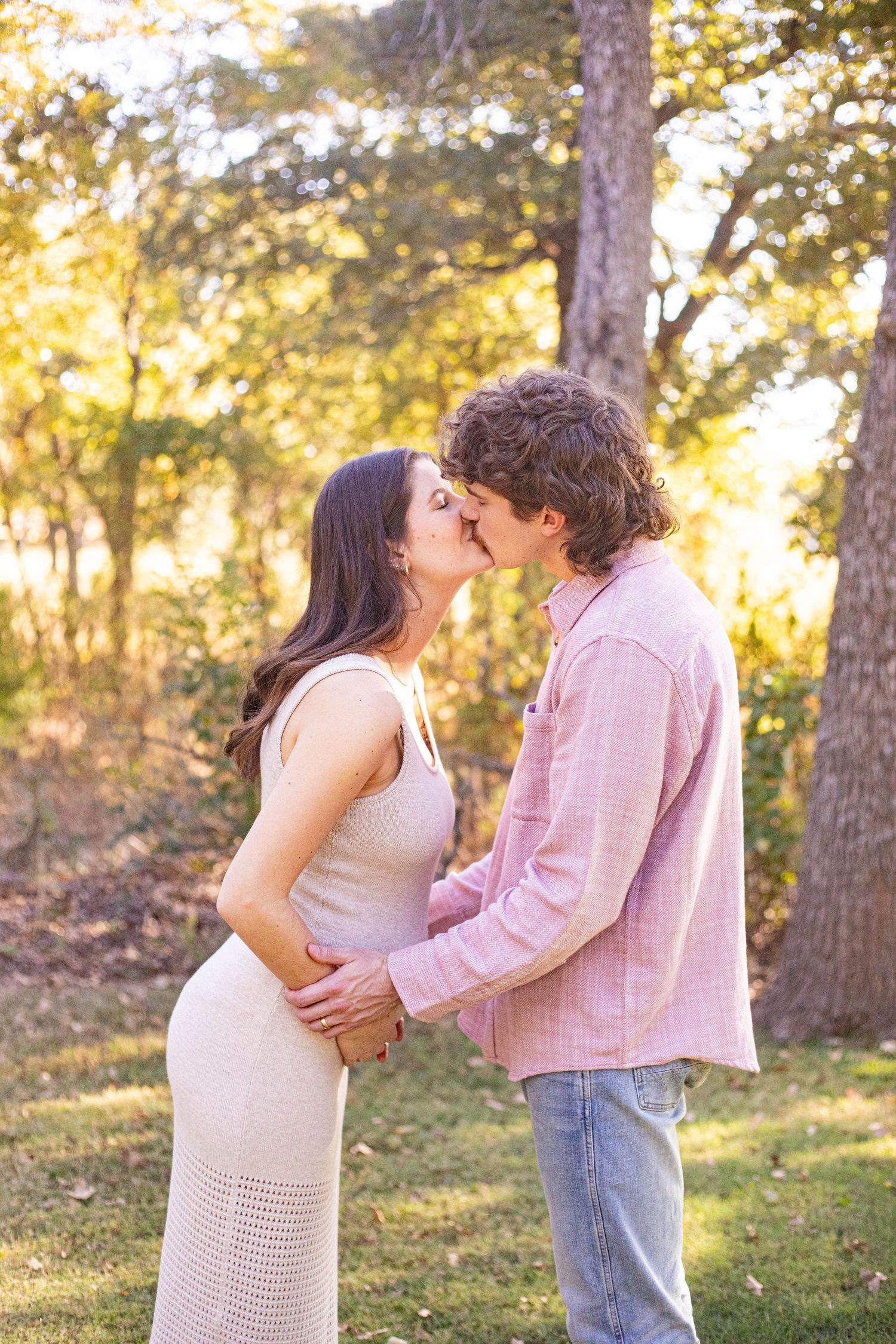 A couple shares a romantic kiss outdoors in a sunlit forest setting, woman in cream dress, man in pink shirt.