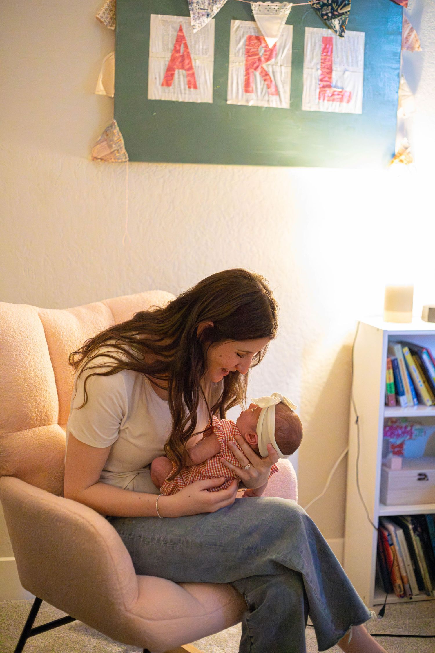 A mother with long brown hair sits in a pink chair cradling her newborn baby in a nursery with colorful letter decorations.